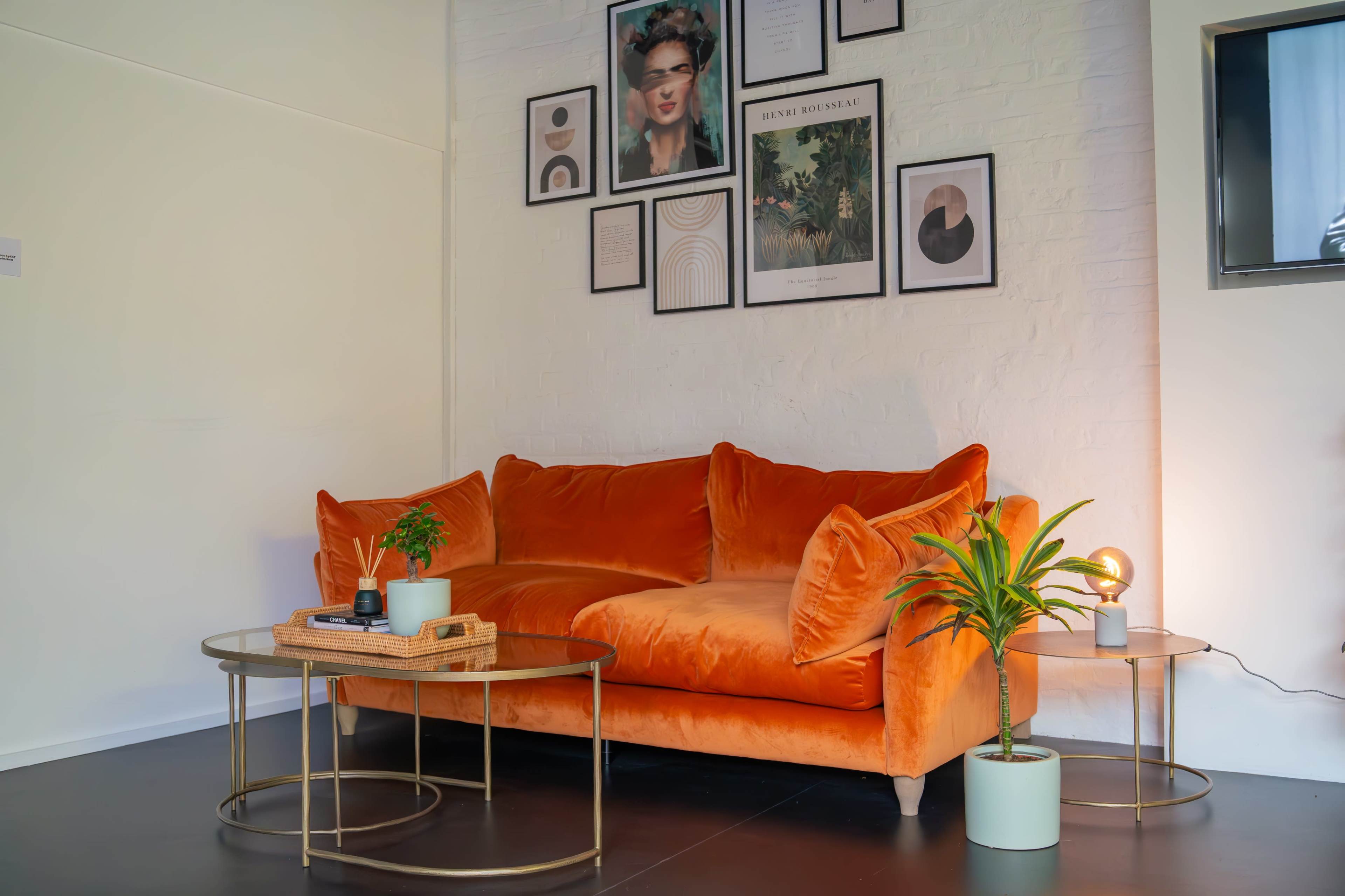 An orange velvet sofa with decorative pillows, flanked by two side tables, a potted plant, and artwork hung on the wall behind it.