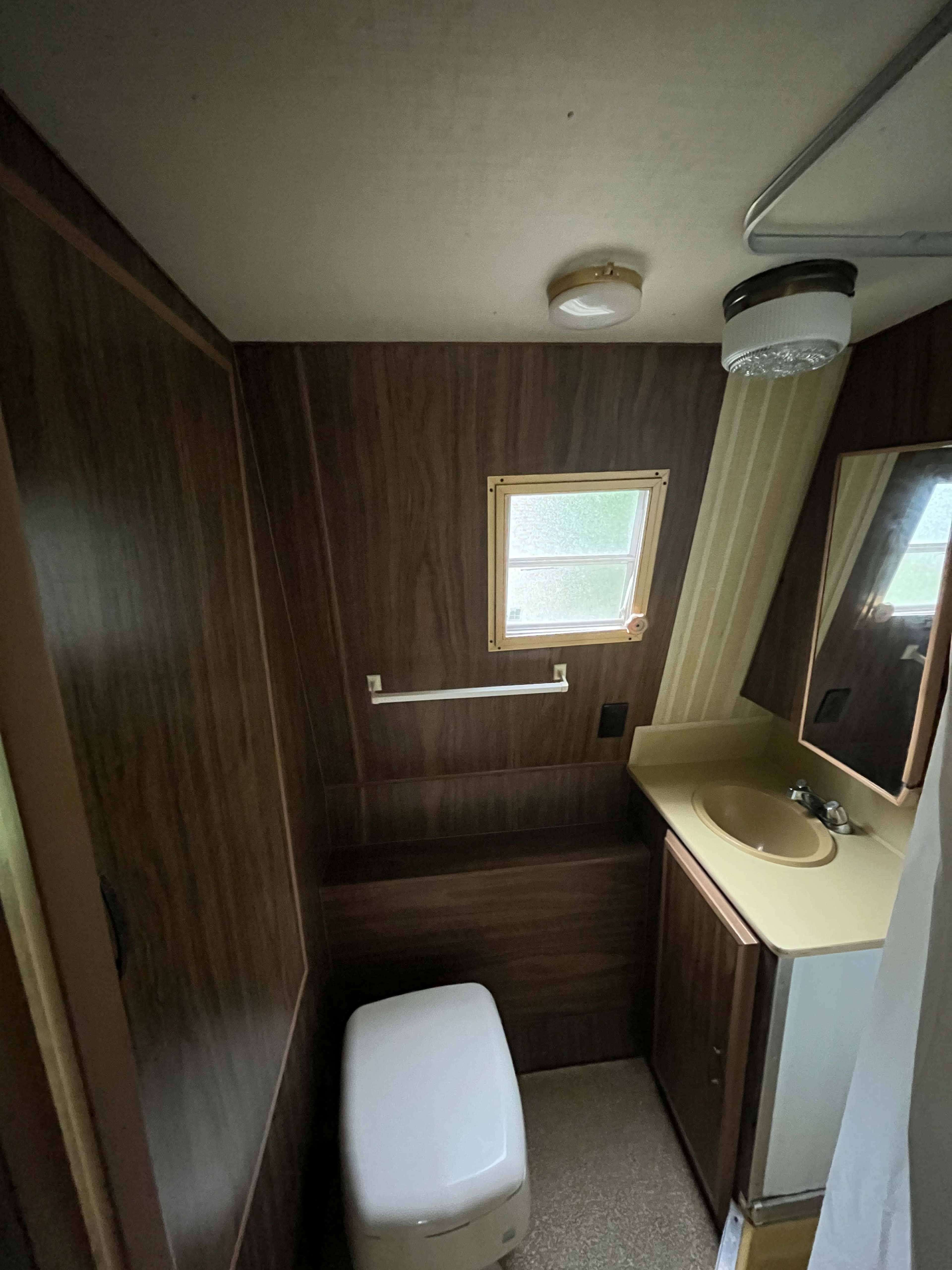 A small, narrow bathroom with wooden paneling, a toilet, a sink, and a window for natural light.