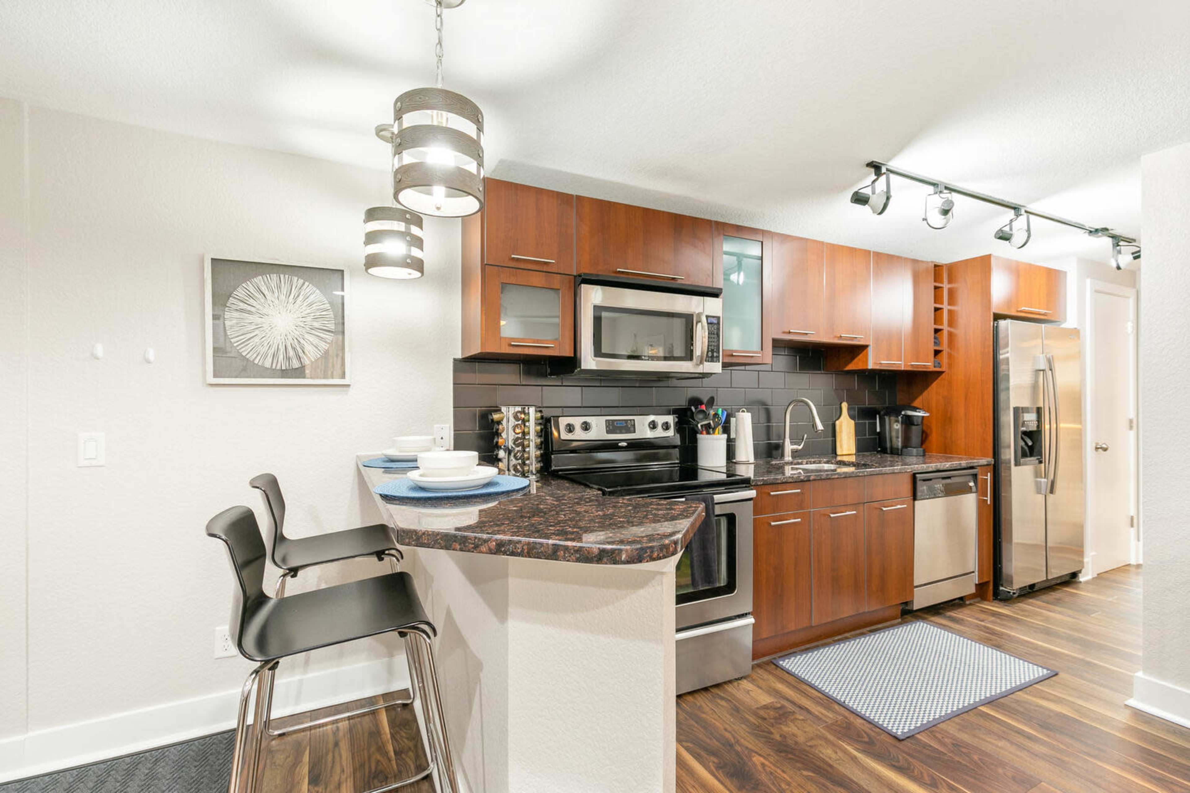 The image shows a modern kitchen with wooden cabinets, stainless steel appliances, and a small breakfast bar with two high chairs.