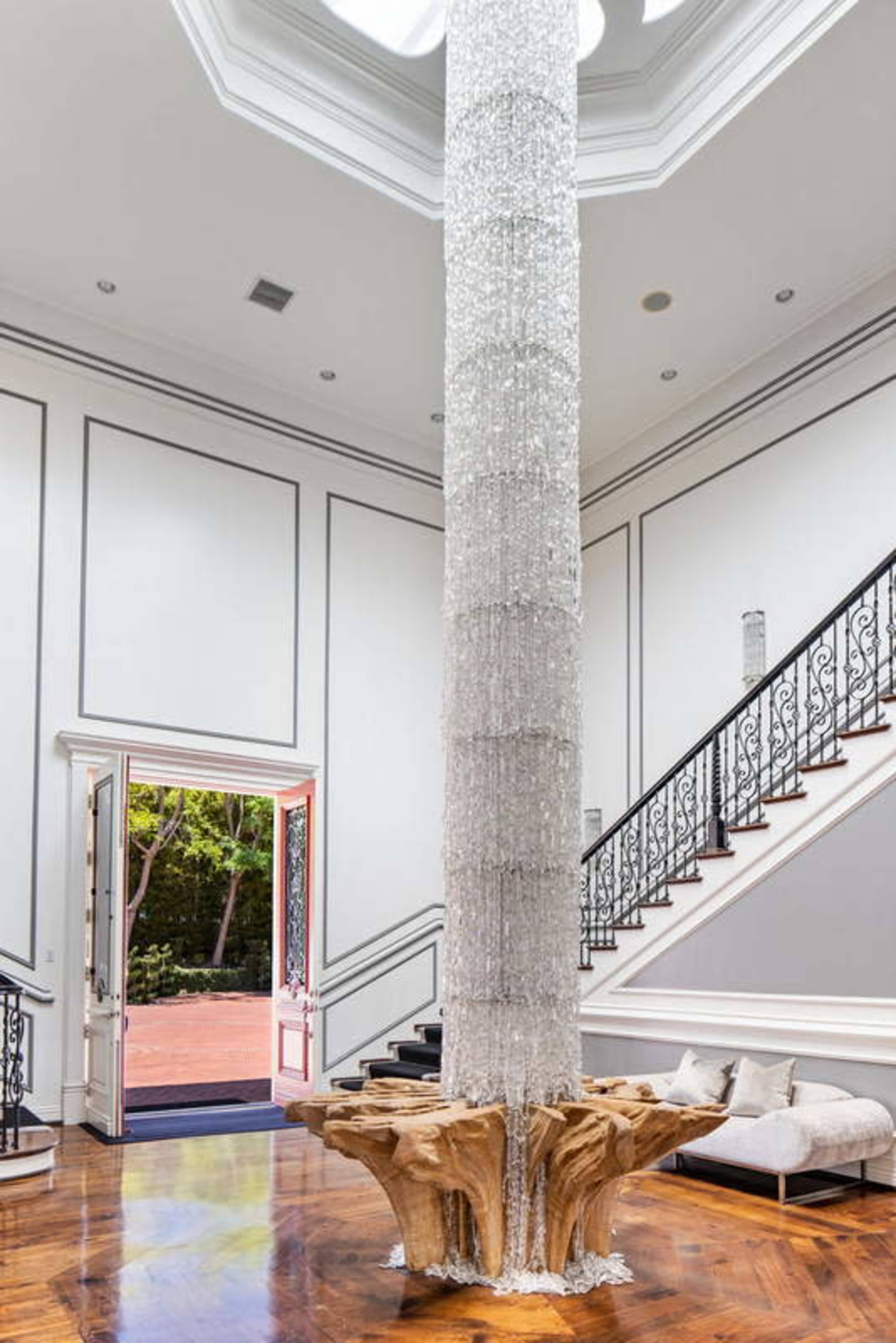 The image features a grand foyer with a tall, crystal chandelier hanging from a circular skylight, accompanied by a unique wooden sculpture base and a staircase with ornate railings.