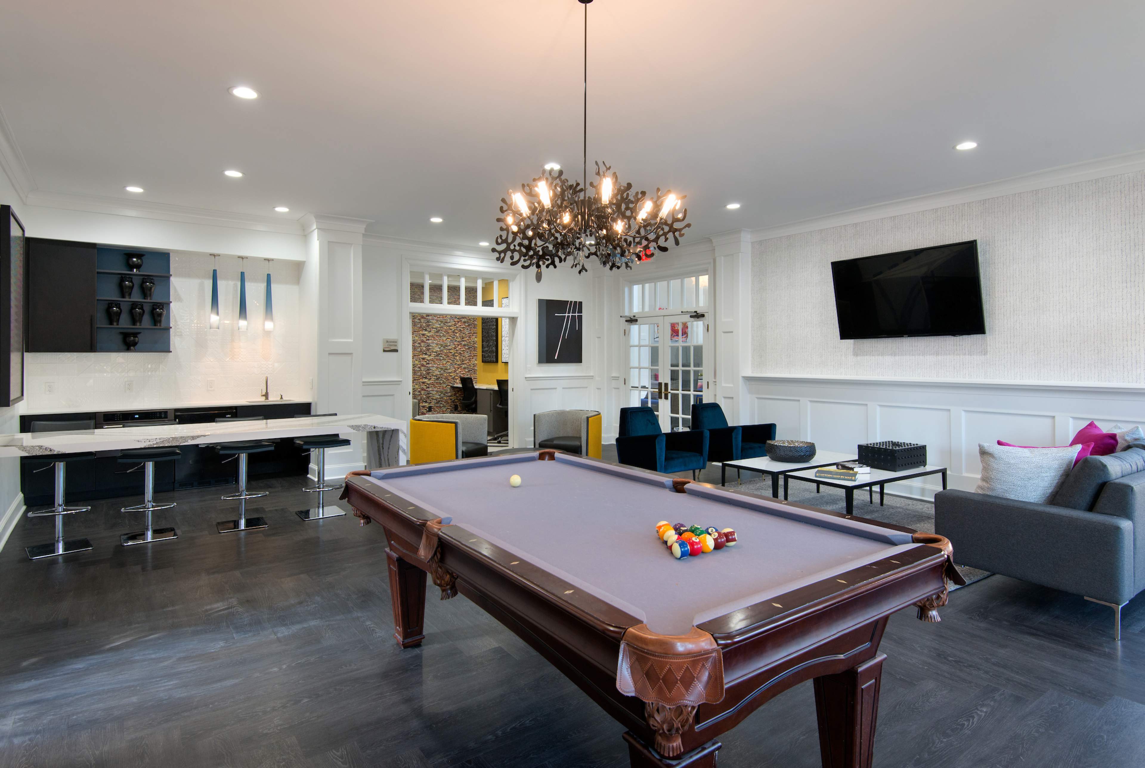 The image shows a modern game room featuring a billiard table, a sleek kitchen counter with bar stools, and comfortable seating areas with a chandelier above.