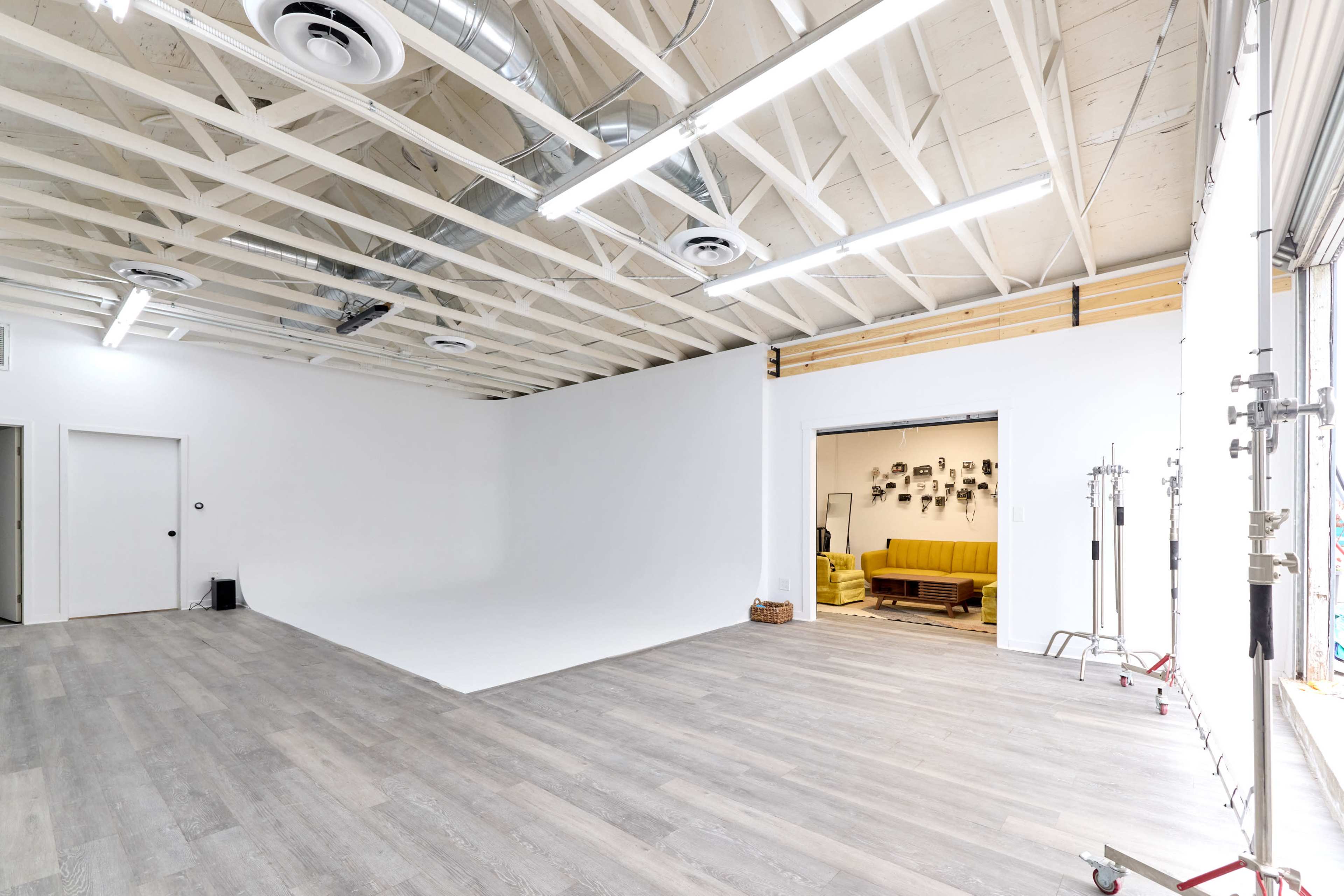 A spacious, well-lit studio with a white backdrop, wooden flooring, and an open entrance leading to a room with a sofa.