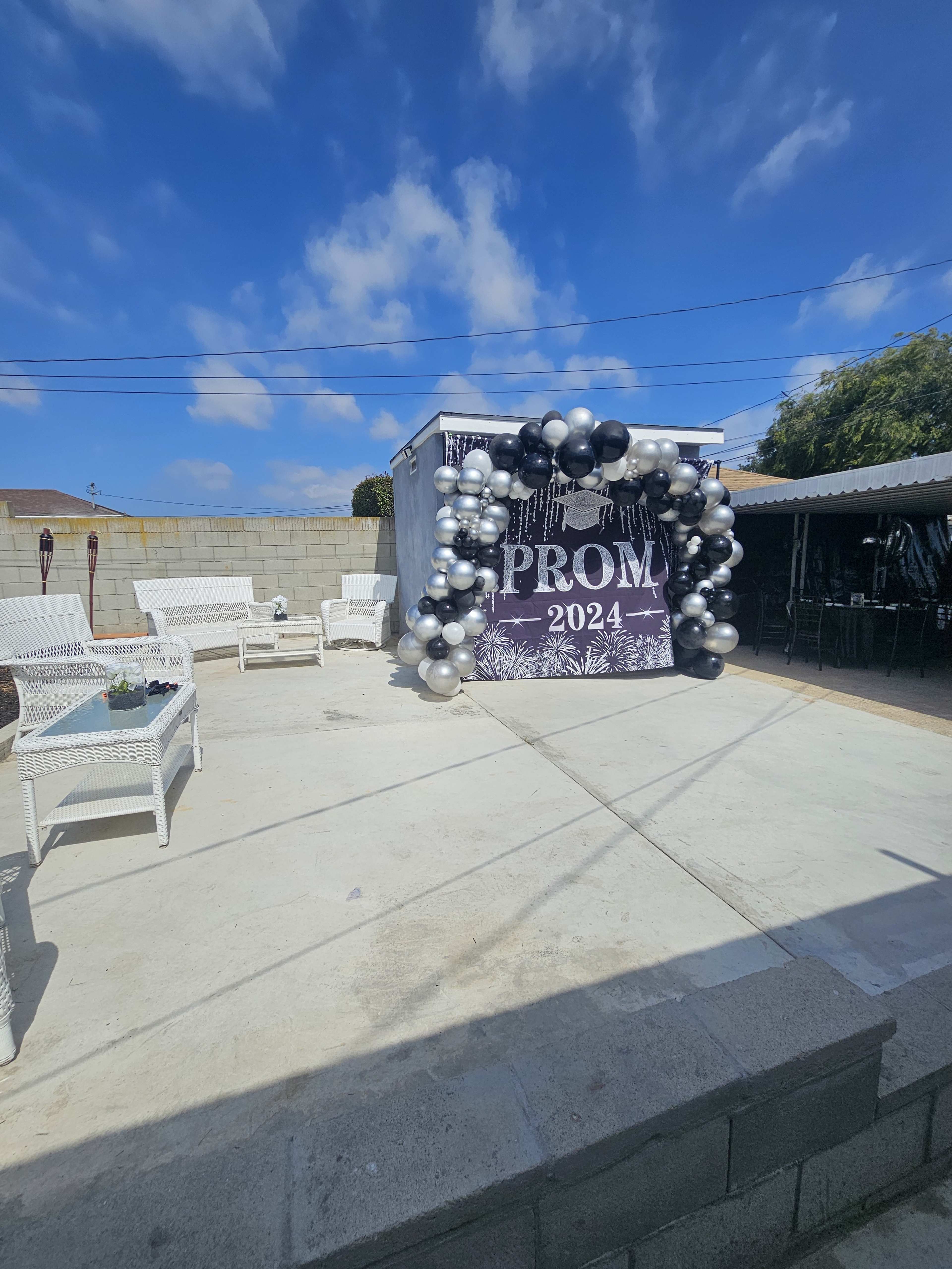 A sign reading "PROM 2024" is framed by a balloon arch in a decorated outdoor space with white furniture and blue skies.