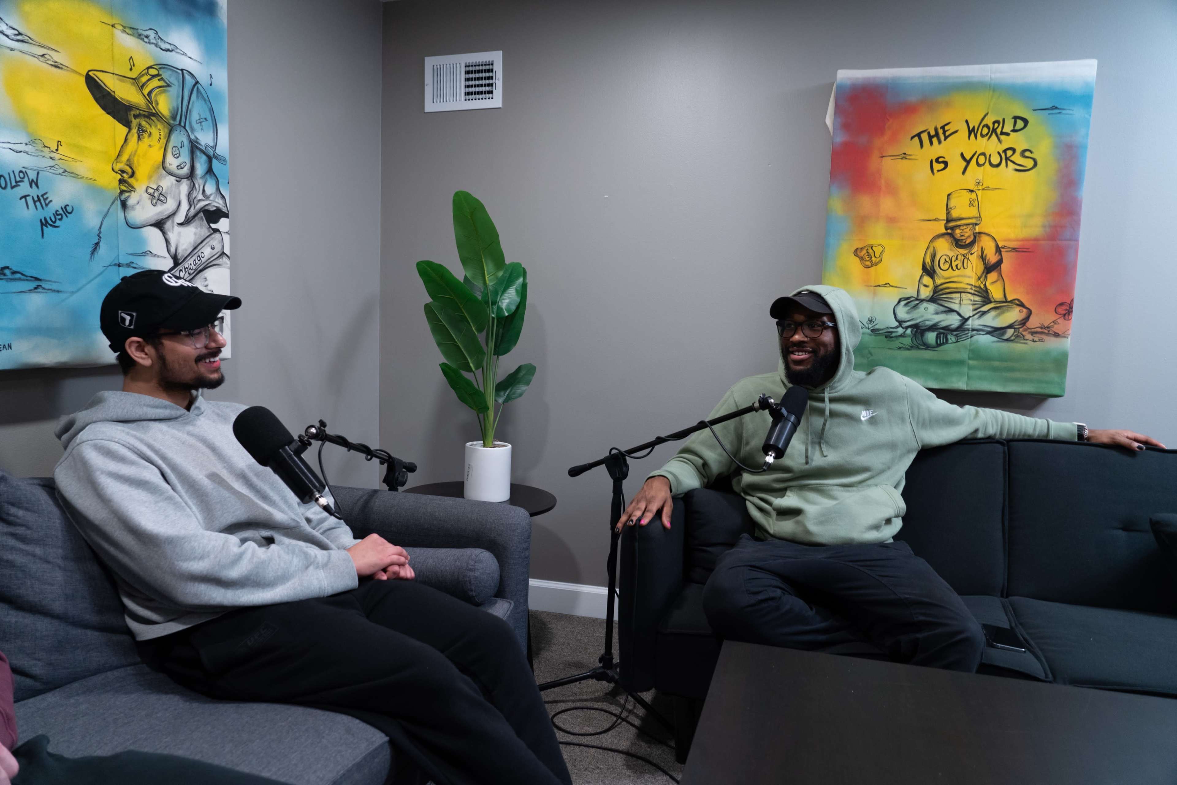 Two men are seated on gray couches in a room with a plant and artwork on the wall, engaged in conversation, with microphones in front of them.