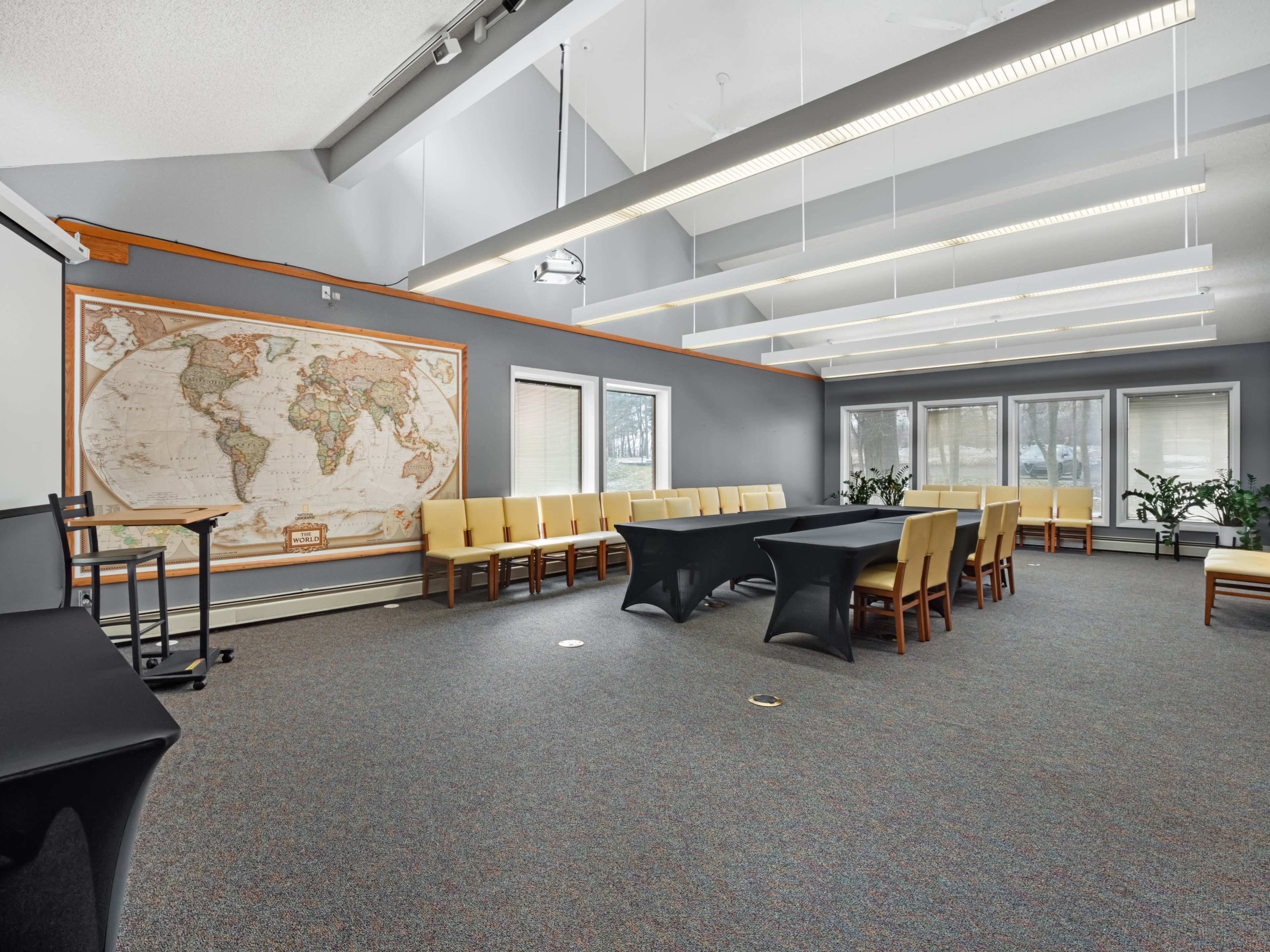 A well-lit conference room featuring a large world map on the wall, a central black table surrounded by chairs, and windows allowing natural light.