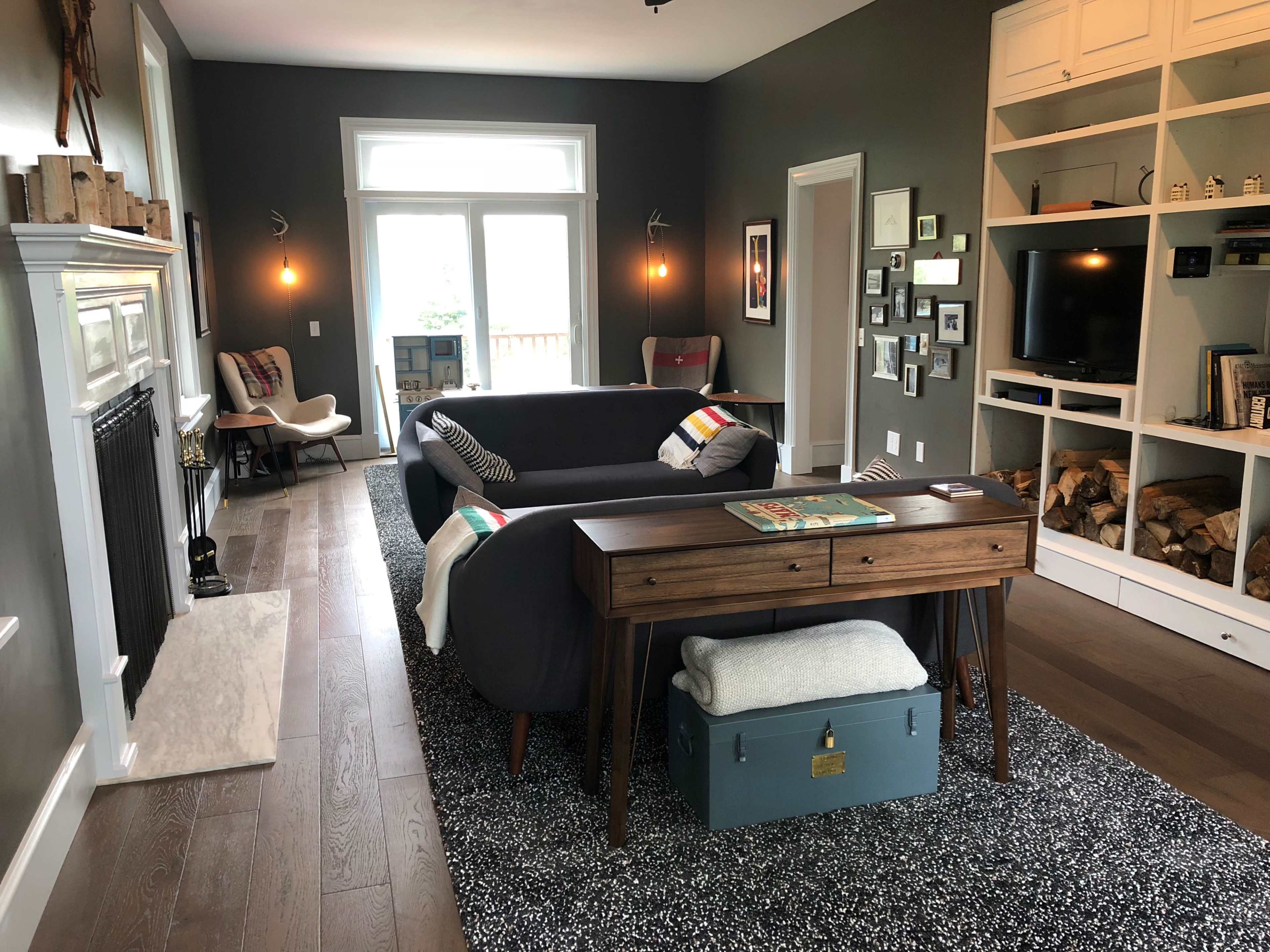 A modern living room with a dark gray wall, a sectional sofa, a wooden console table, and a shelving unit filled with decorative items, alongside a window and a door leading to a patio.