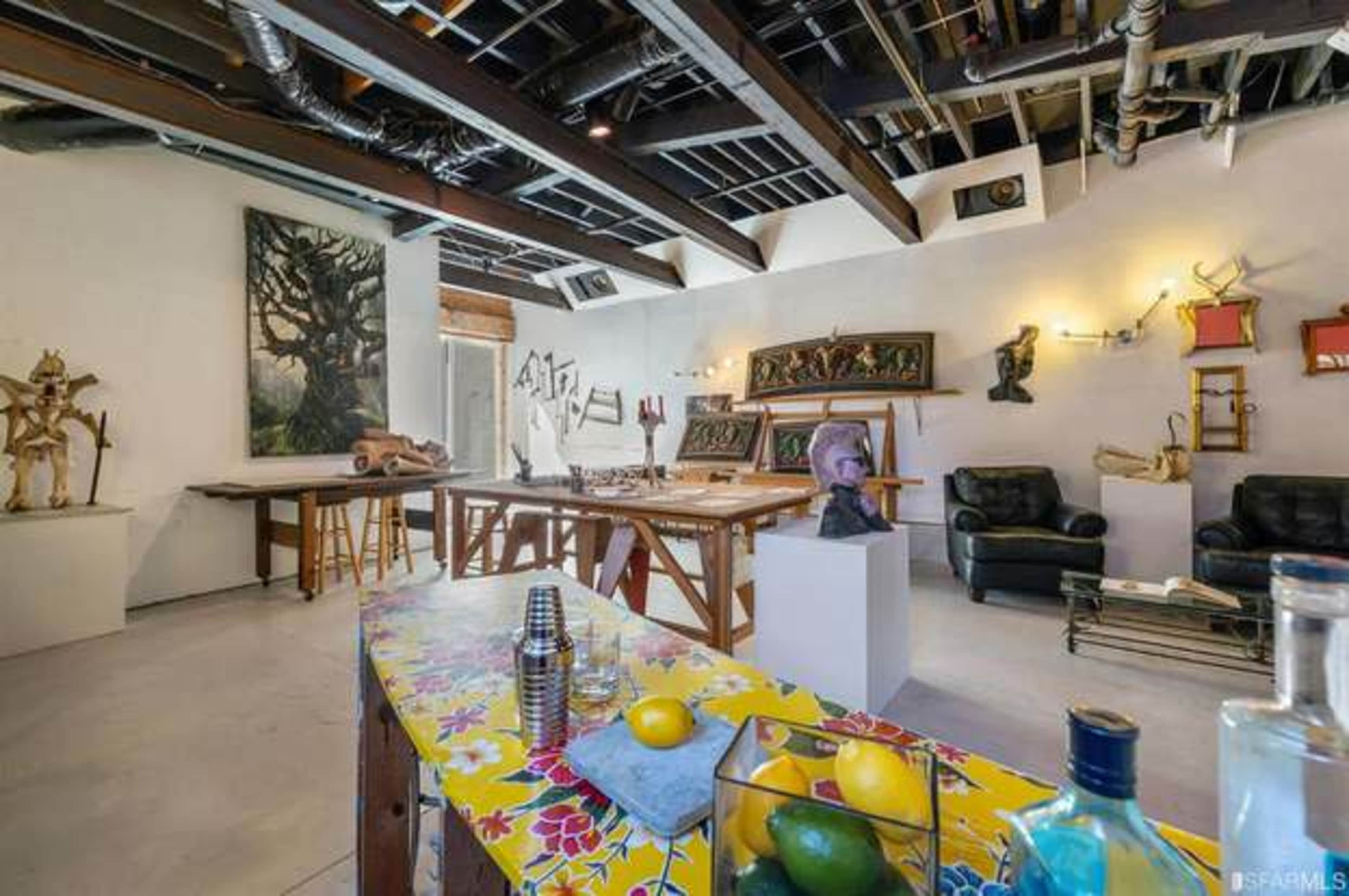 The image shows a spacious art studio featuring various tools, sculptures, and artwork, with a colorful table in the foreground and seating arrangements in the background.