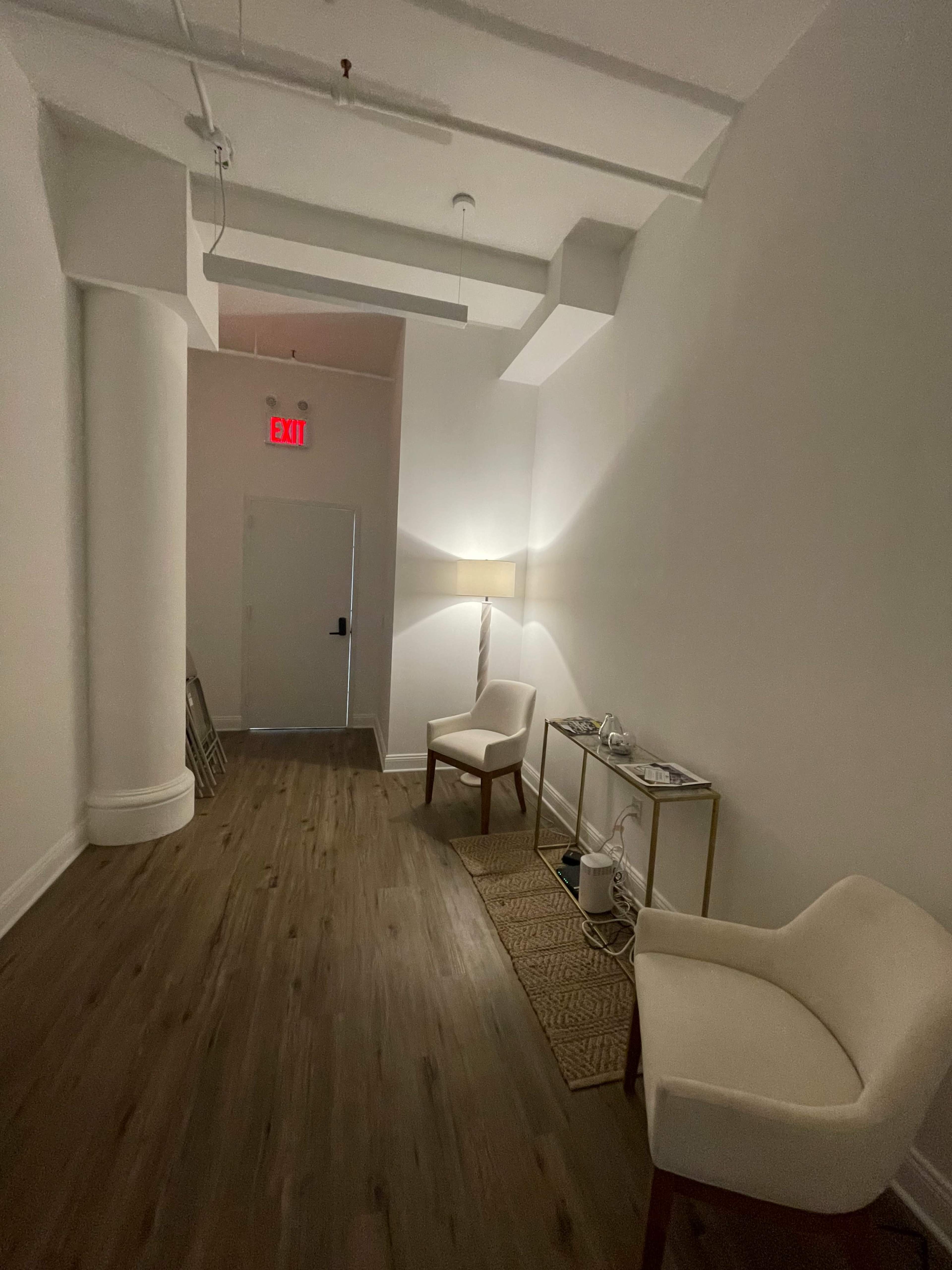 A hallway with white walls, wooden flooring, two chairs, a console table with items, and an exit door at the end.