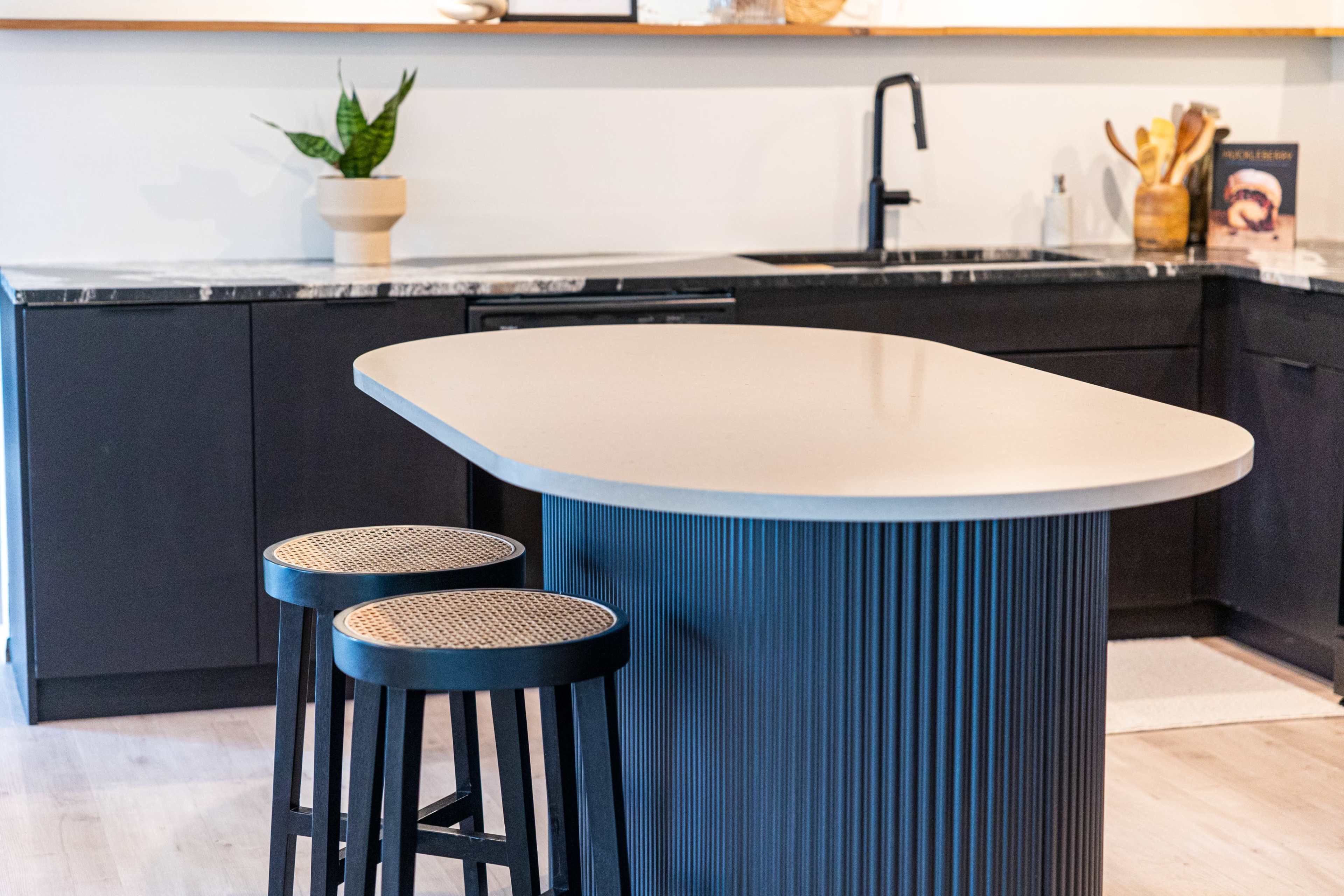 A modern kitchen features a curved island with two wooden stools, dark cabinetry, and a sink with a minimalist faucet.