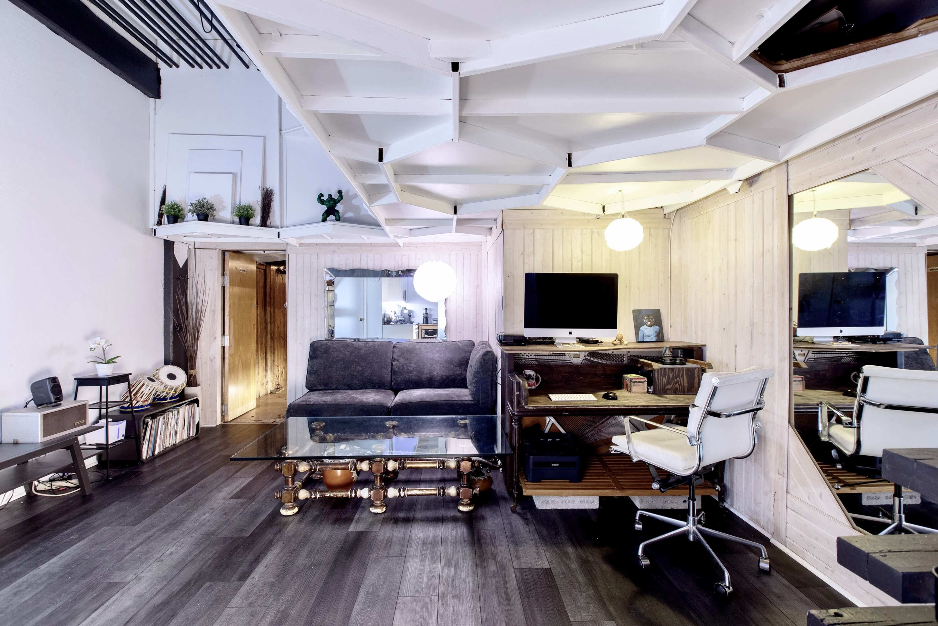 The image shows a modern living room with a glass coffee table, a gray sofa, a desk with a computer, and mirrored walls.