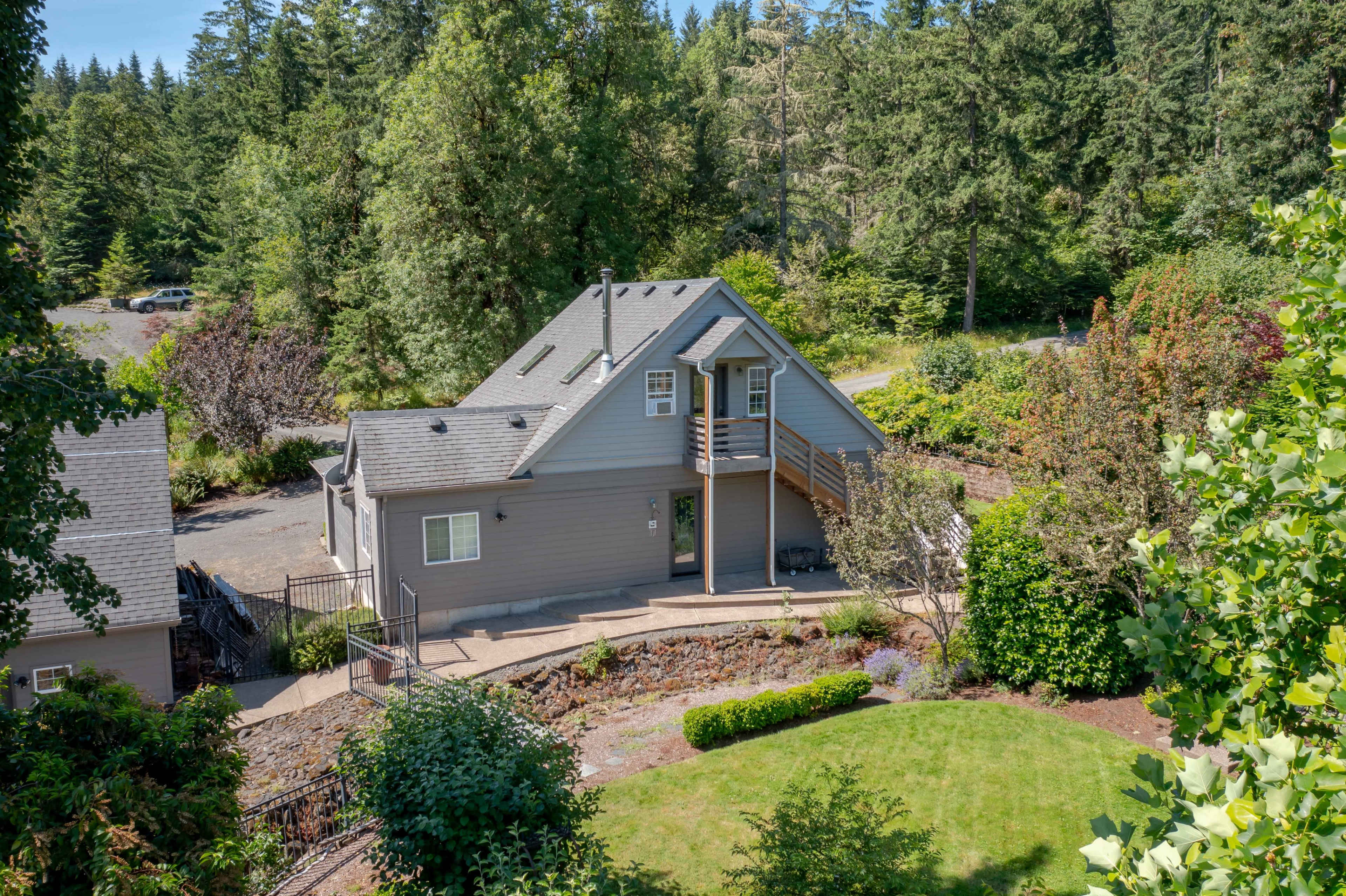 A two-story gray house surrounded by greenery and trees with a gravel driveway visible in the background.