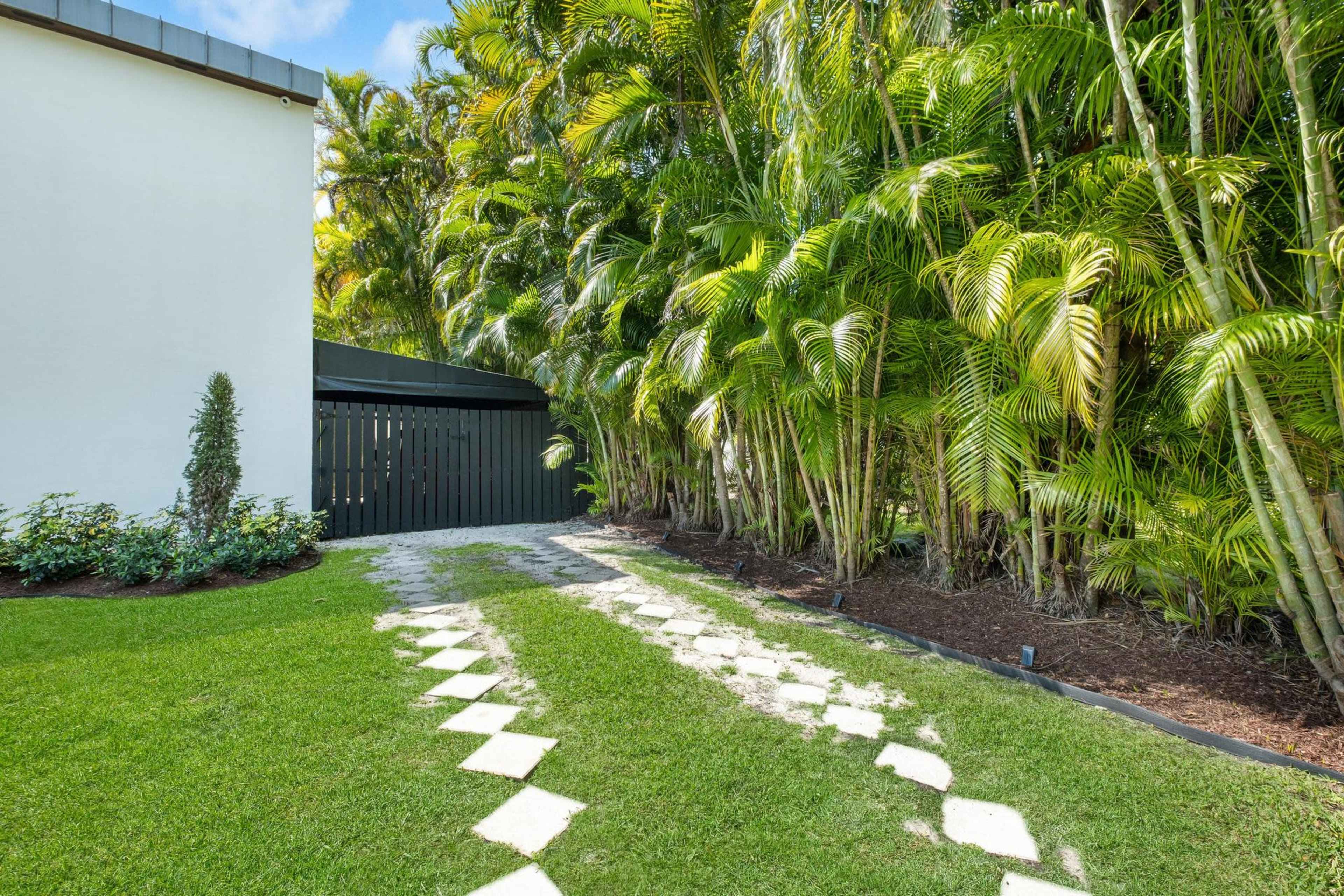 A pathway of stepping stones leads through lush greenery next to a fence and a white building.