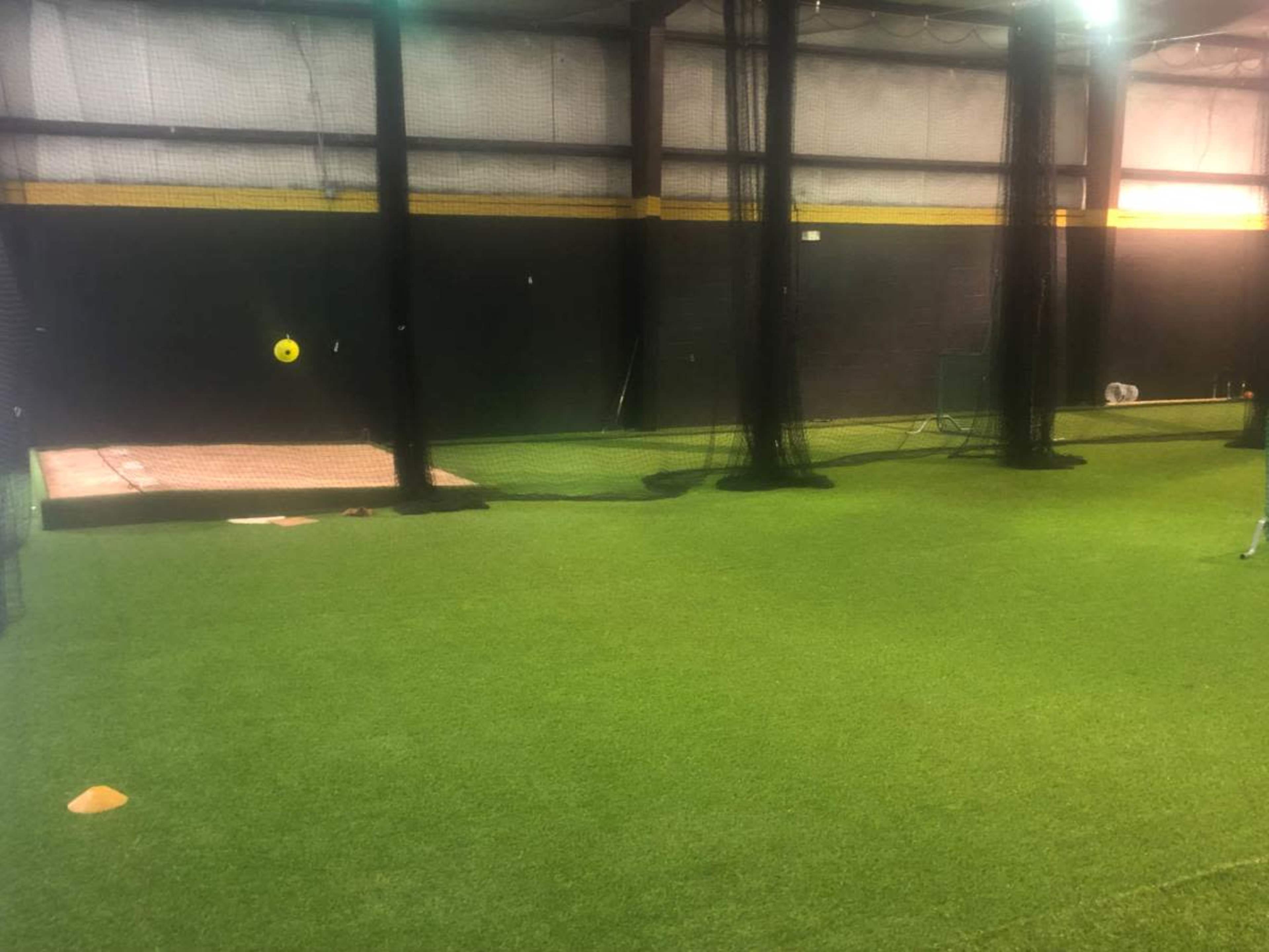 The image shows an indoor sports facility with artificial turf, batting cages on the left, and a pitching area in the back.
