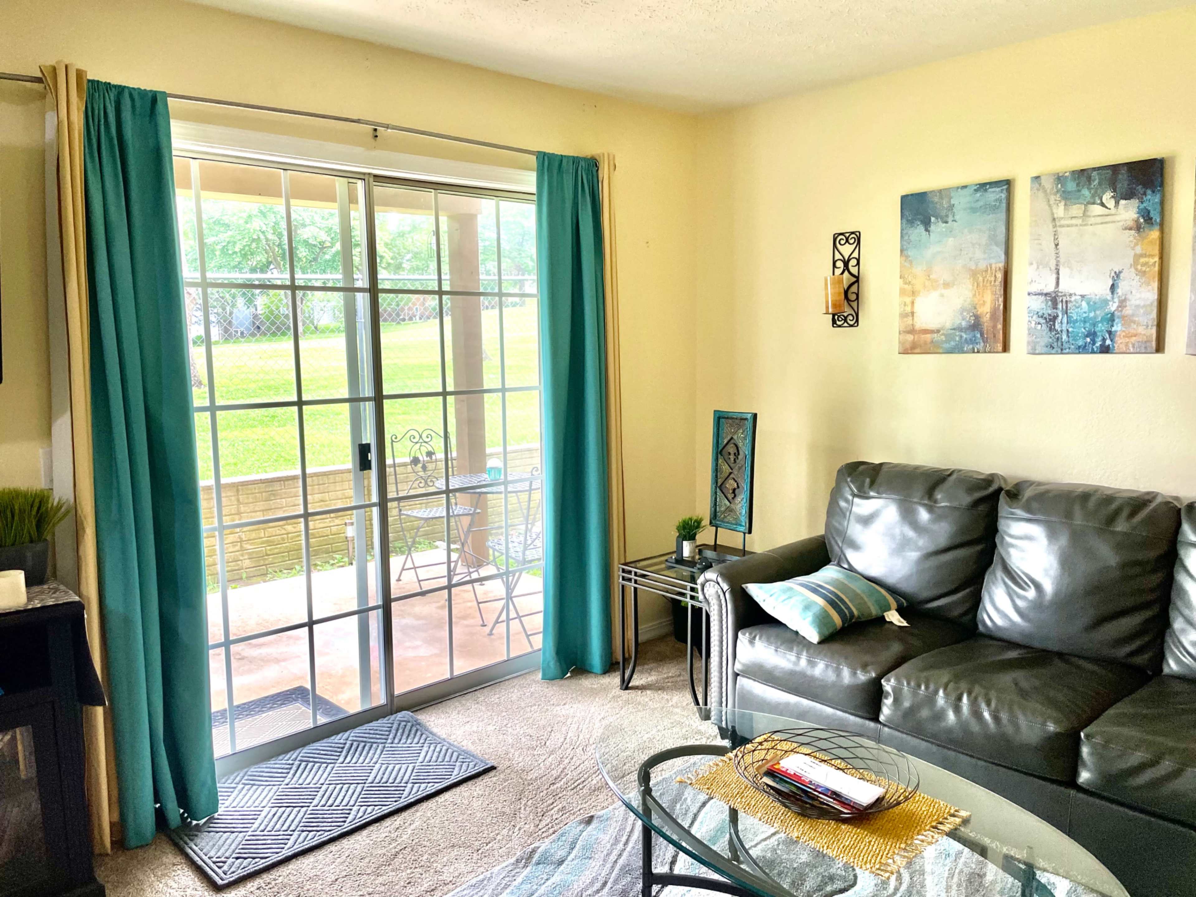 A living room features a gray sofa facing a glass coffee table, with a sliding door leading to a patio and two abstract paintings on the wall.