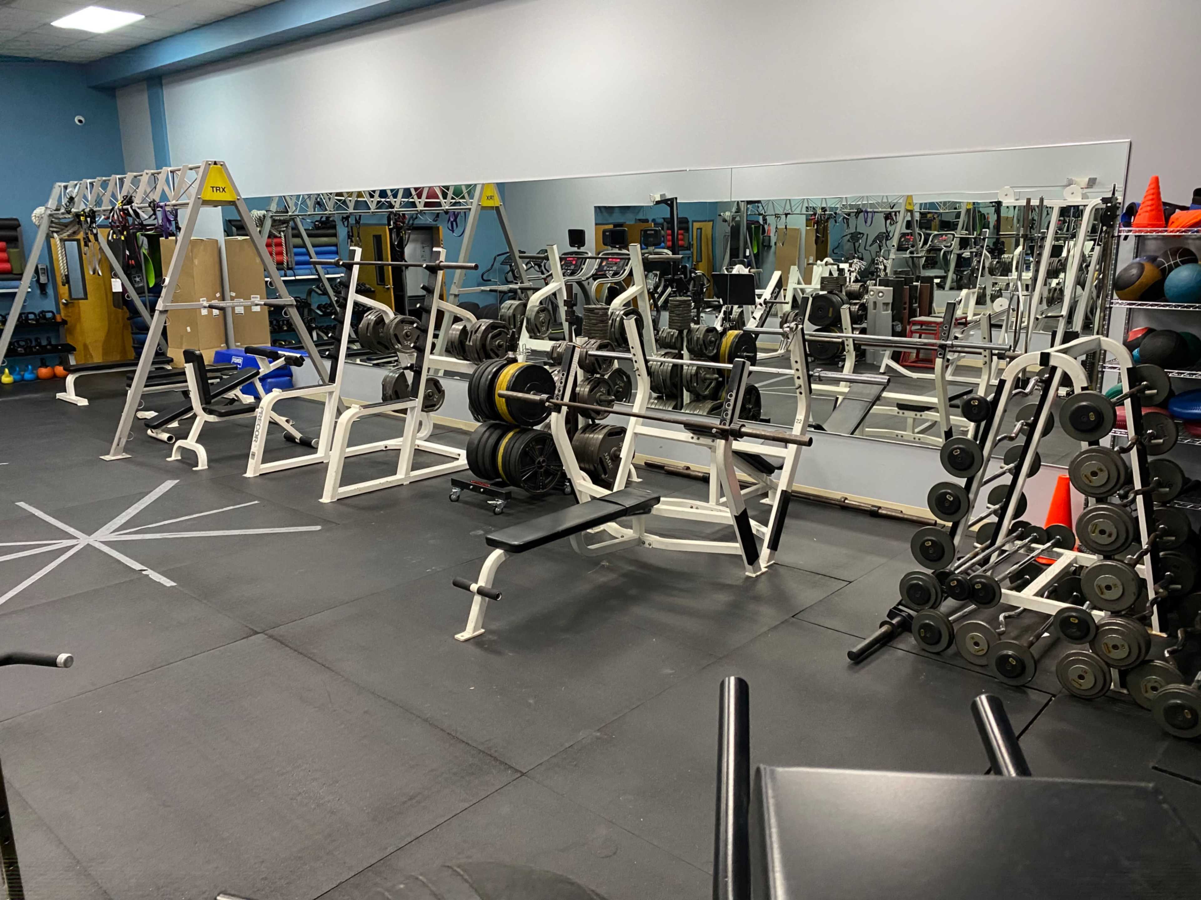A gym with various weightlifting equipment, including benches and a rack of dumbbells, set against a mirrored wall.