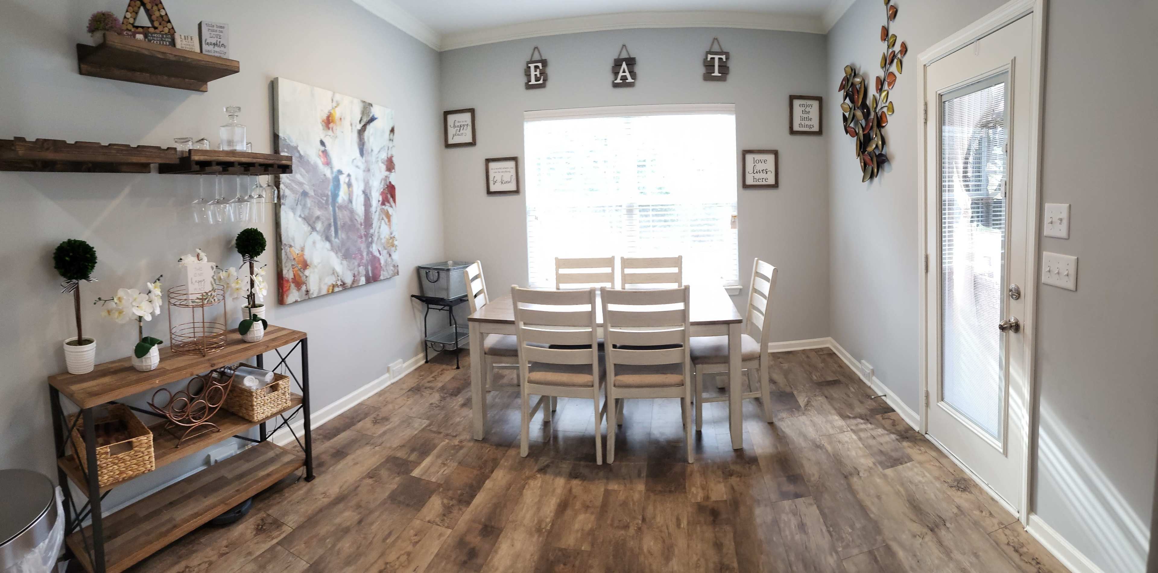 A dining area features a wooden table with white chairs, a shelf with decorative items, and a large window with blinds.