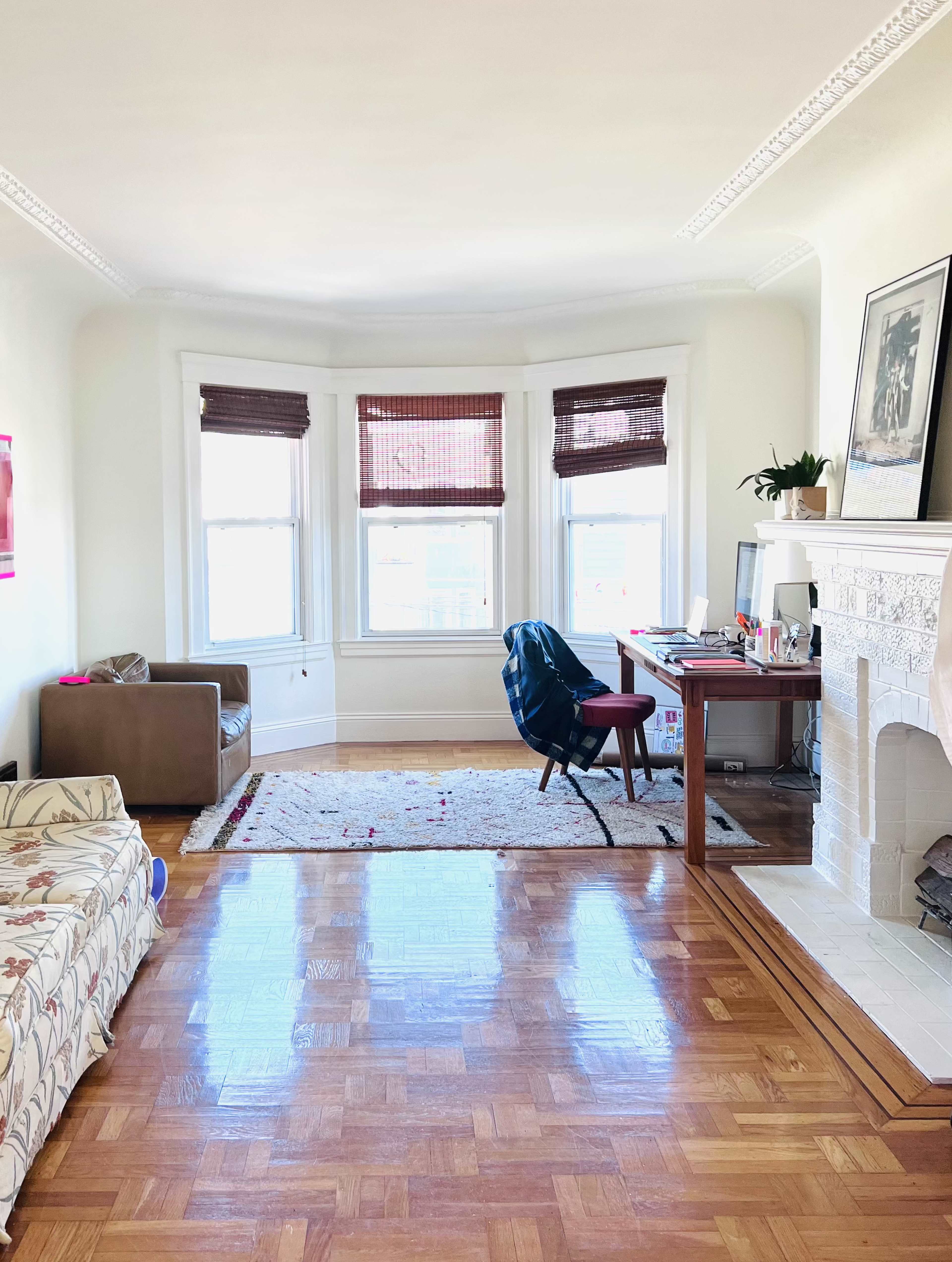 A tidy living room features a corner sofa, a chair with a desk and computer, and large windows with wooden blinds.