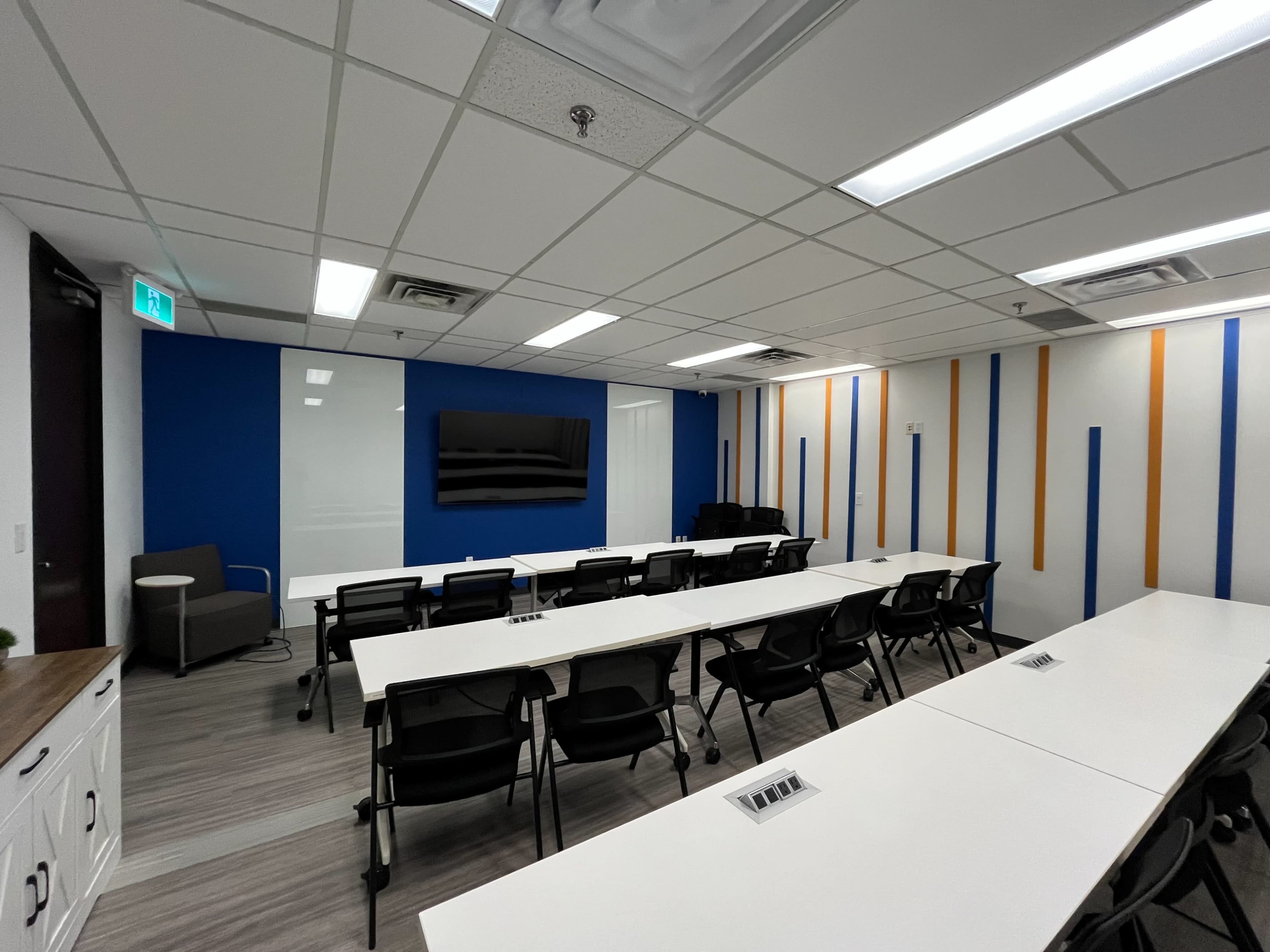 The image shows a modern conference room with several rows of tables and chairs arranged facing a wall-mounted television.