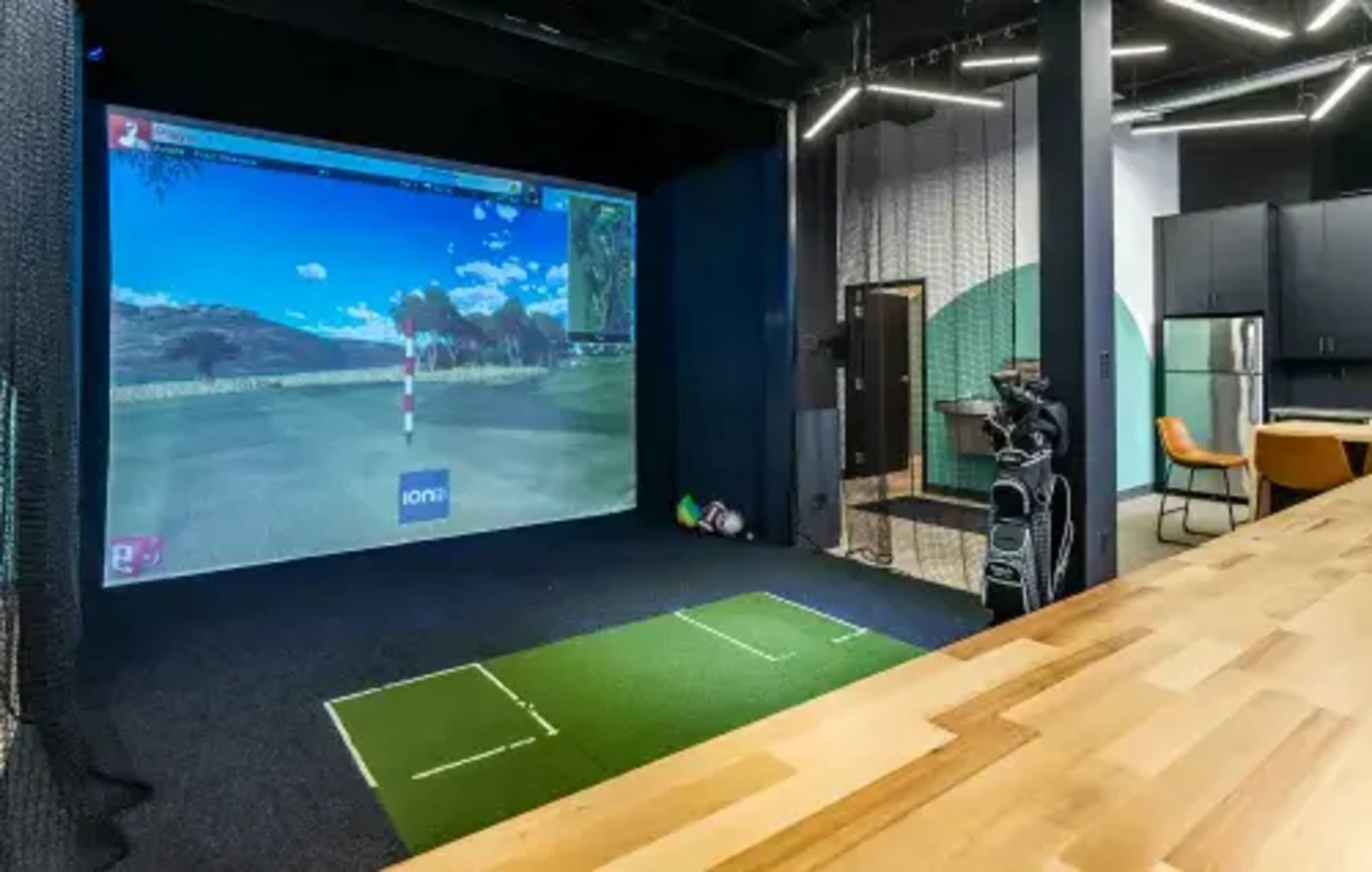 A virtual golf simulator setup featuring a large projection screen, a putting green, and golf clubs arranged nearby.