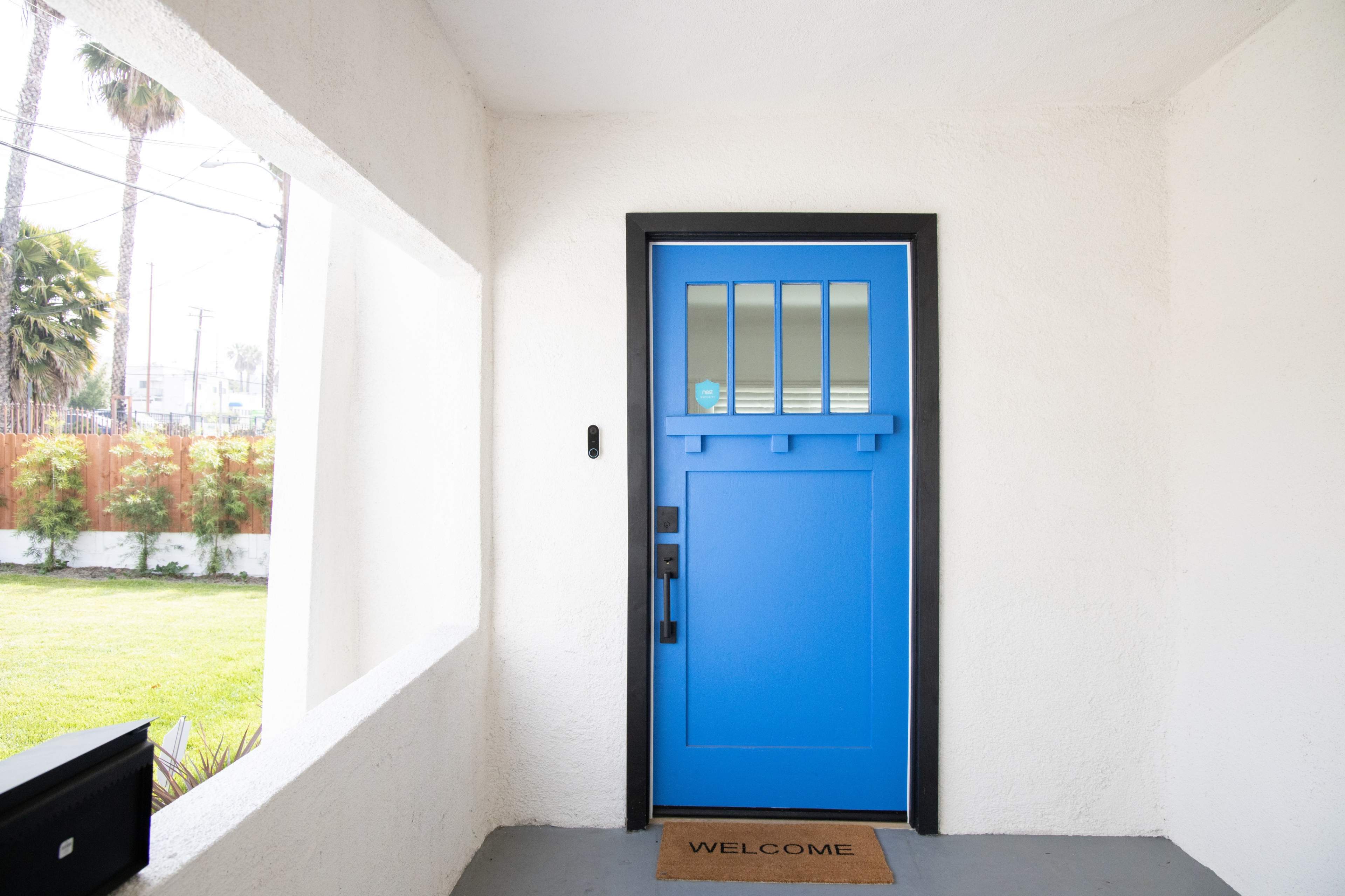 A blue front door with a small window is framed by a white stucco wall, with a welcome mat on the floor and a grassy area visible outside.