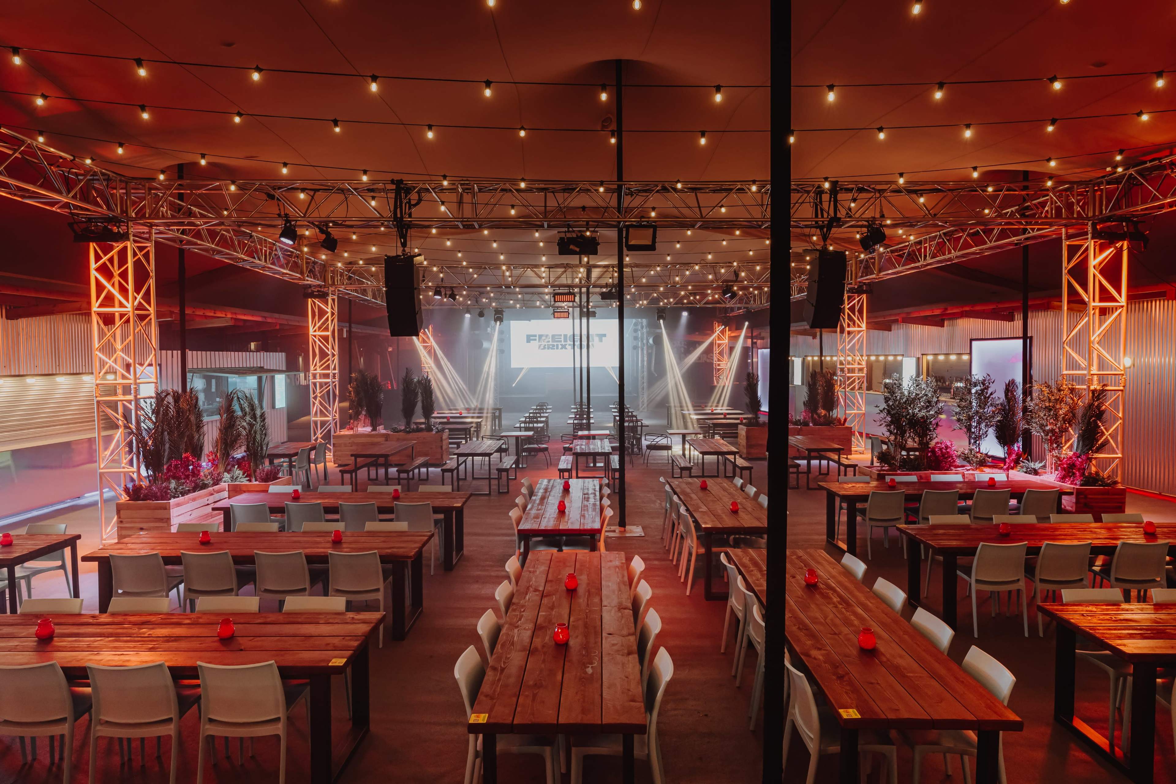 A large event space features long wooden tables set for dining, surrounded by bright lights and a stage at the front.