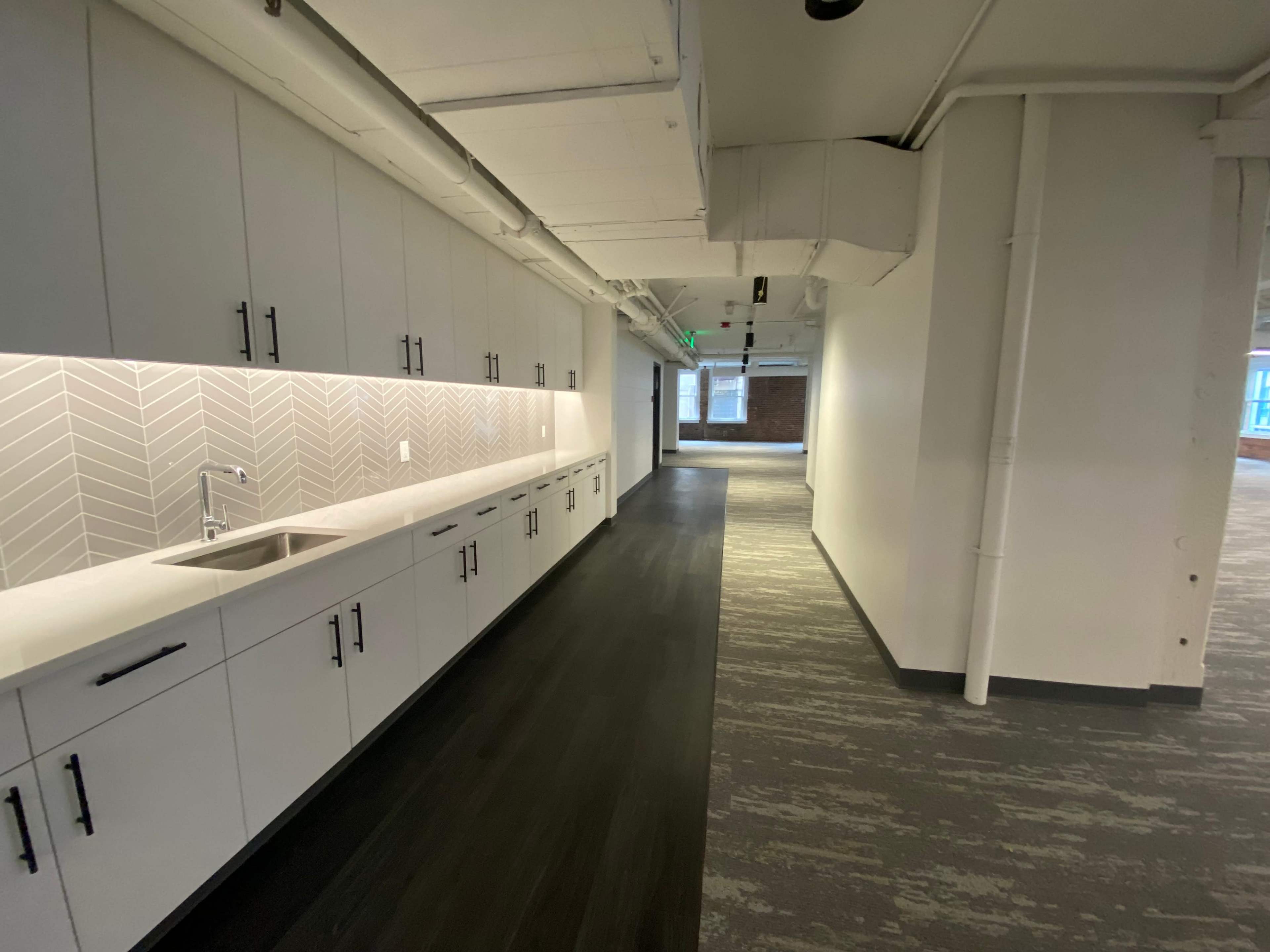 The image shows a modern kitchen area with a long countertop, cabinets, and a herringbone-patterned backsplash, situated in a well-lit corridor of an office space.