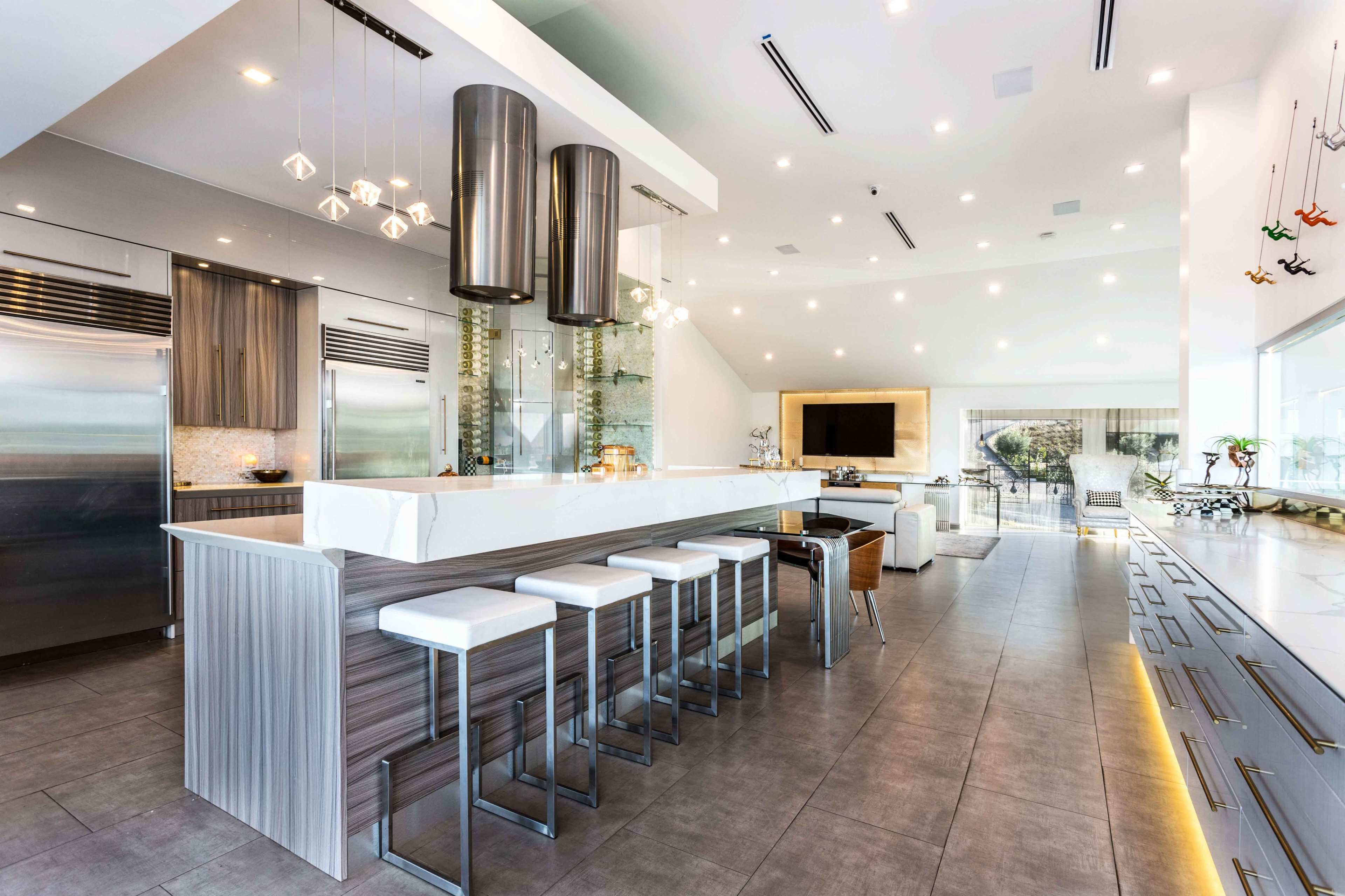 A modern kitchen features a large island with white bar stools, pendant lighting, and stainless steel appliances.