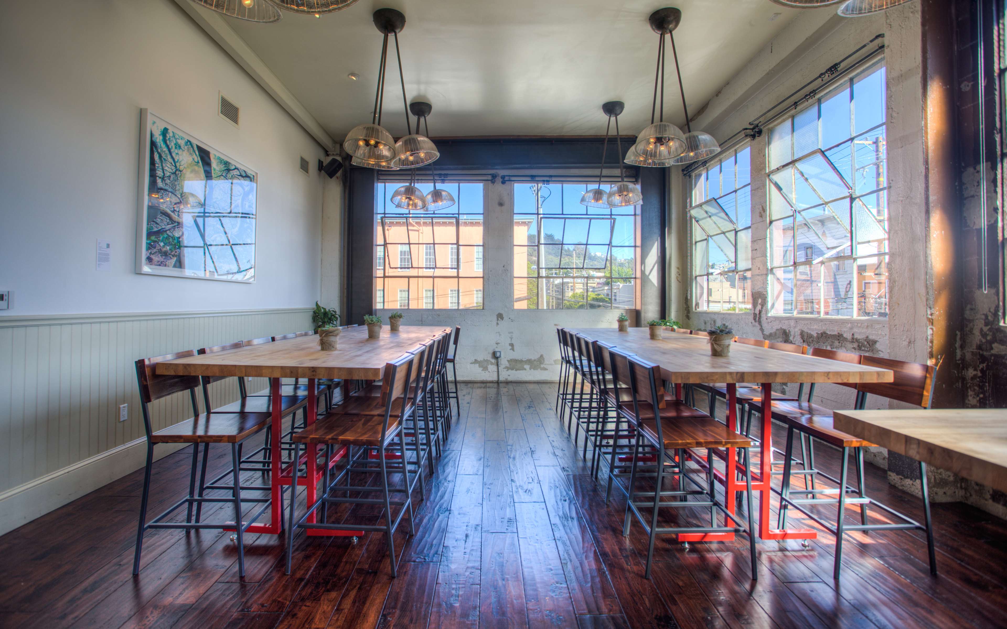 The image shows a spacious dining room with long wooden tables, industrial-style chairs, and large windows allowing natural light to fill the space.