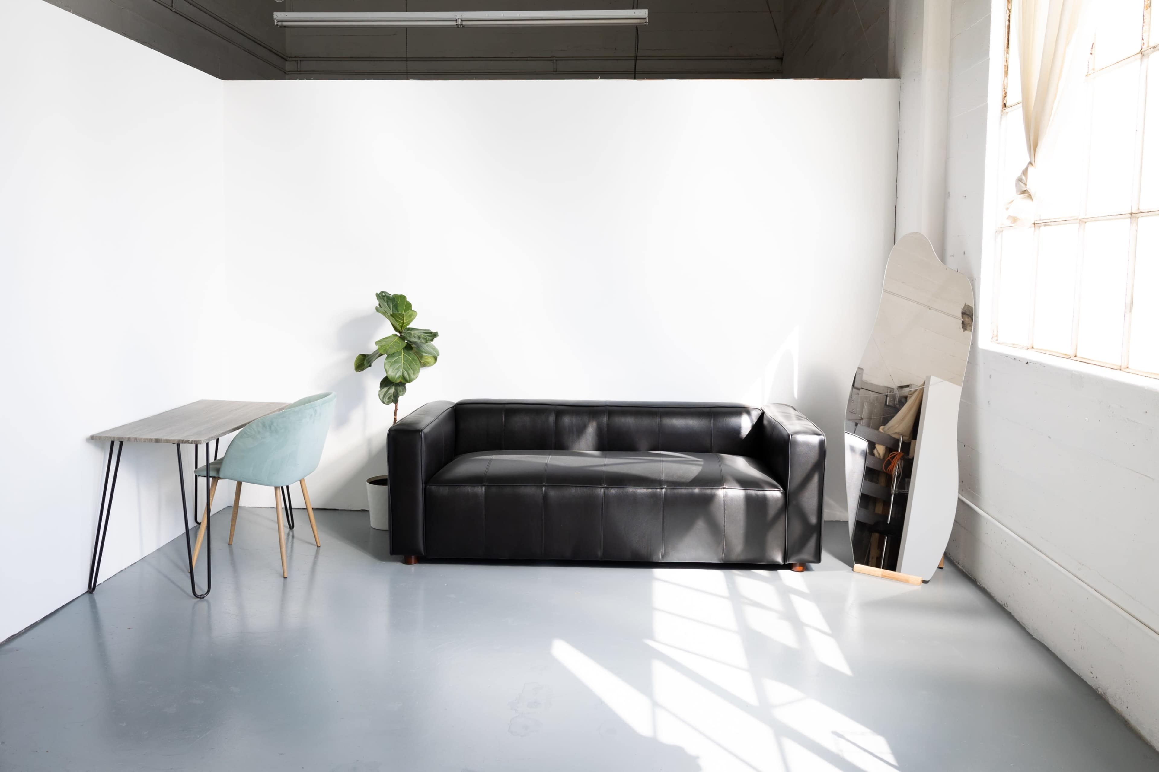 A black leather sofa is positioned against a white wall, accompanied by a small table, a light blue chair, a potted plant, and a surfboard leaning against the wall.