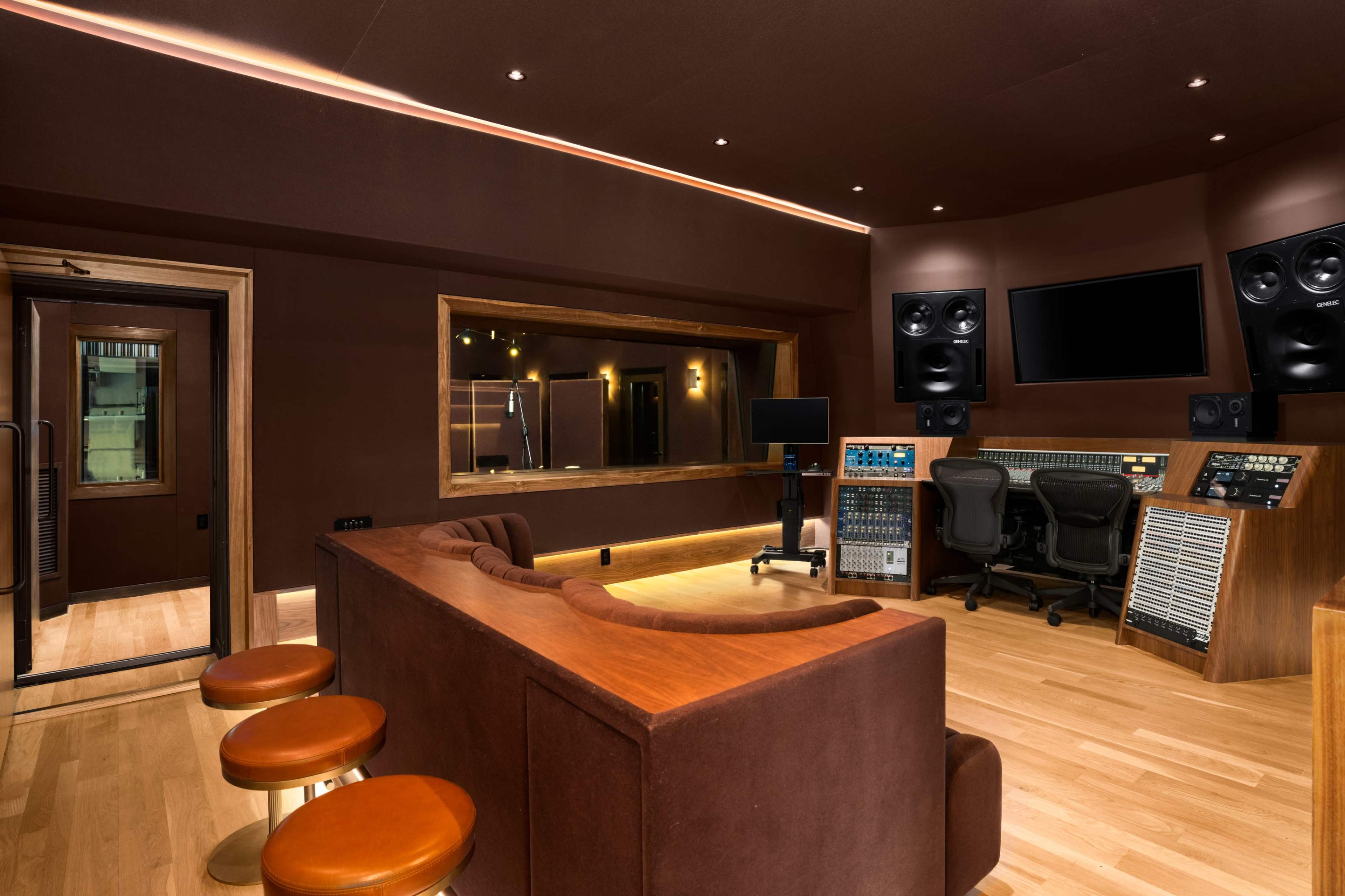 The image shows a sound recording studio with a brown color scheme, featuring a mixing console, large speakers, and a sofa with bar stools arranged around it.