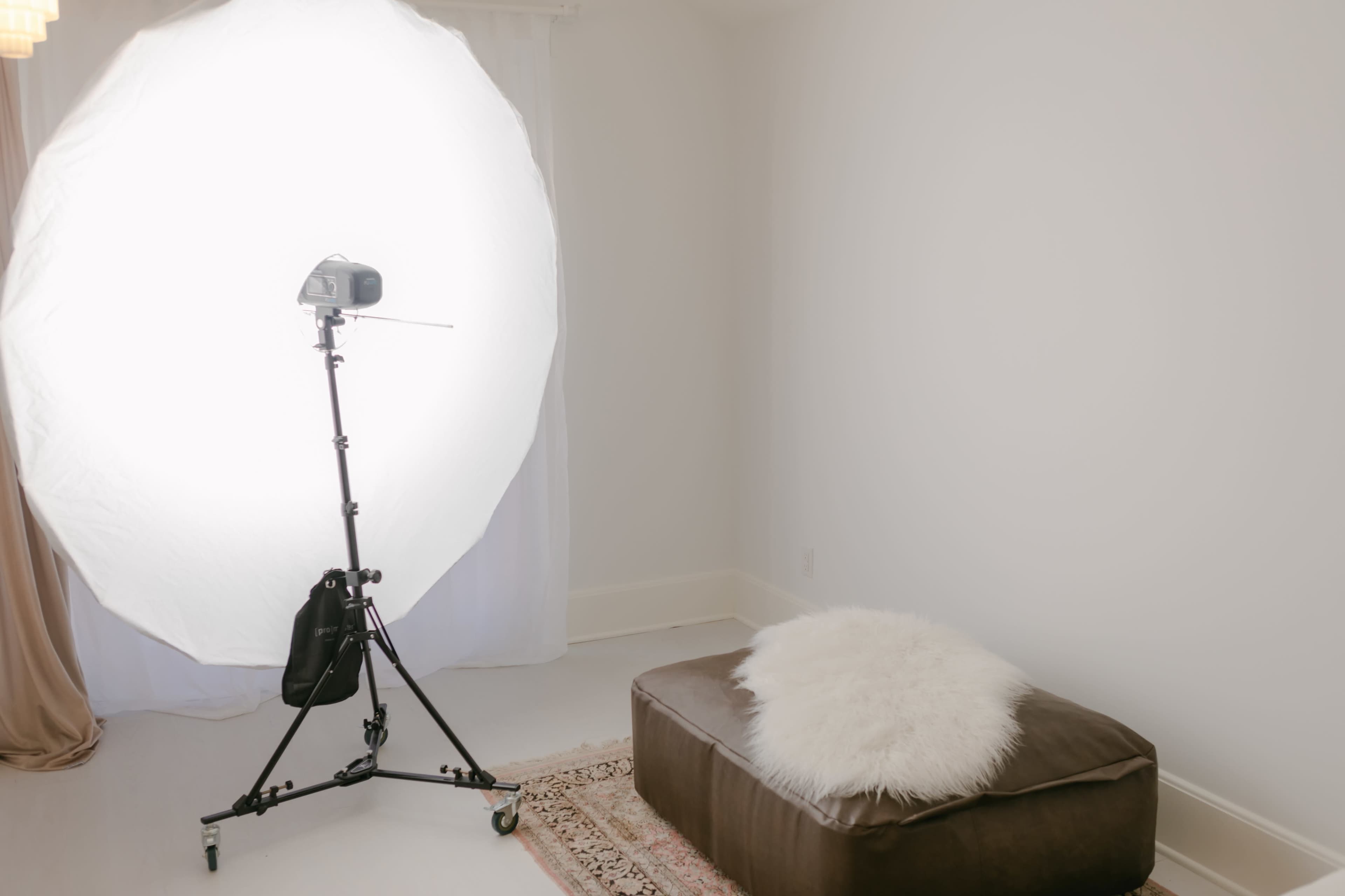 A photography setup features a large softbox light on a stand next to a brown ottoman with a white fur throw on it, in a minimalistic room.