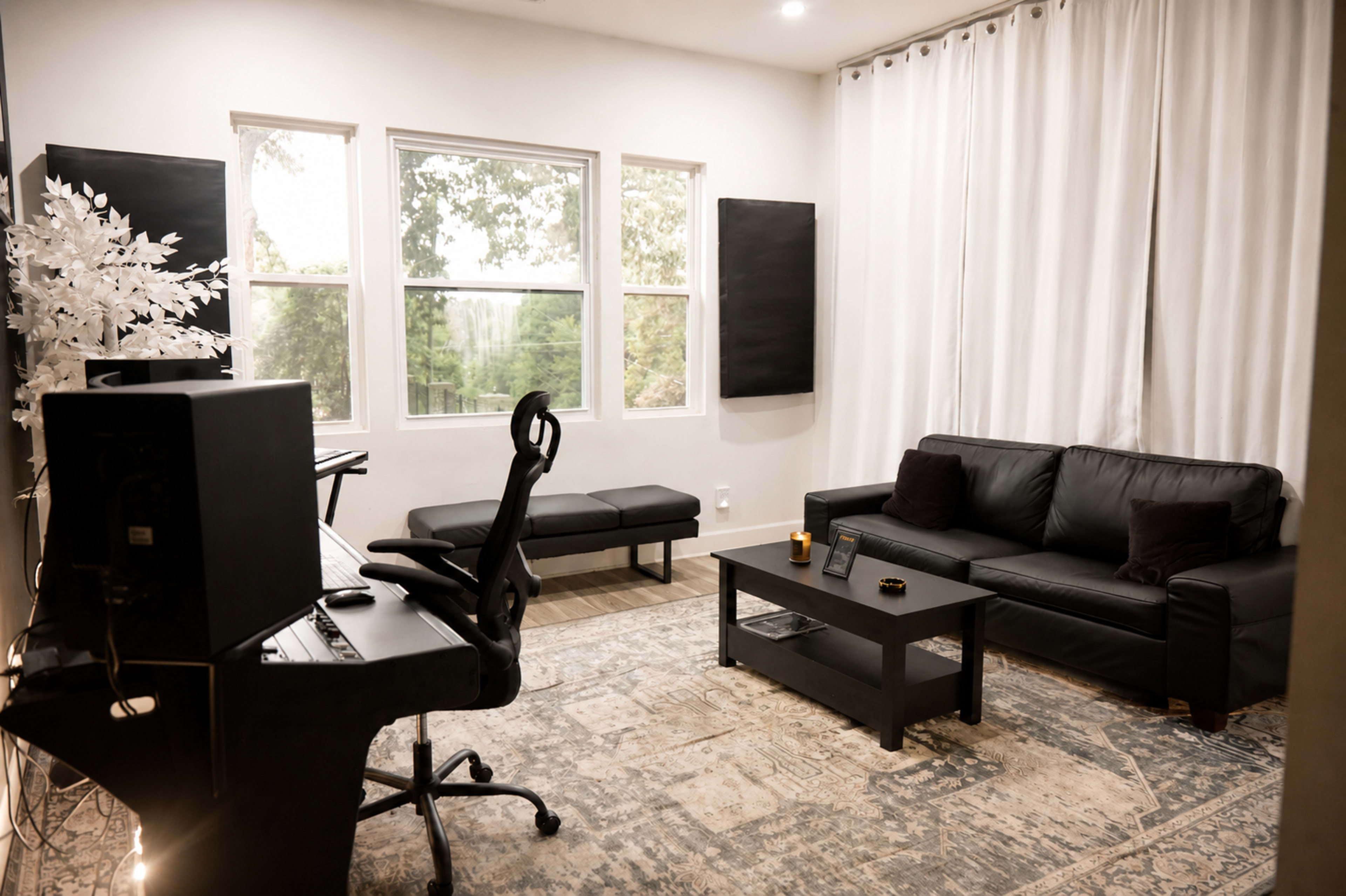 The image shows a modern, minimalistic living room with a black sofa, a coffee table, a workspace with a computer, and large windows allowing natural light in.