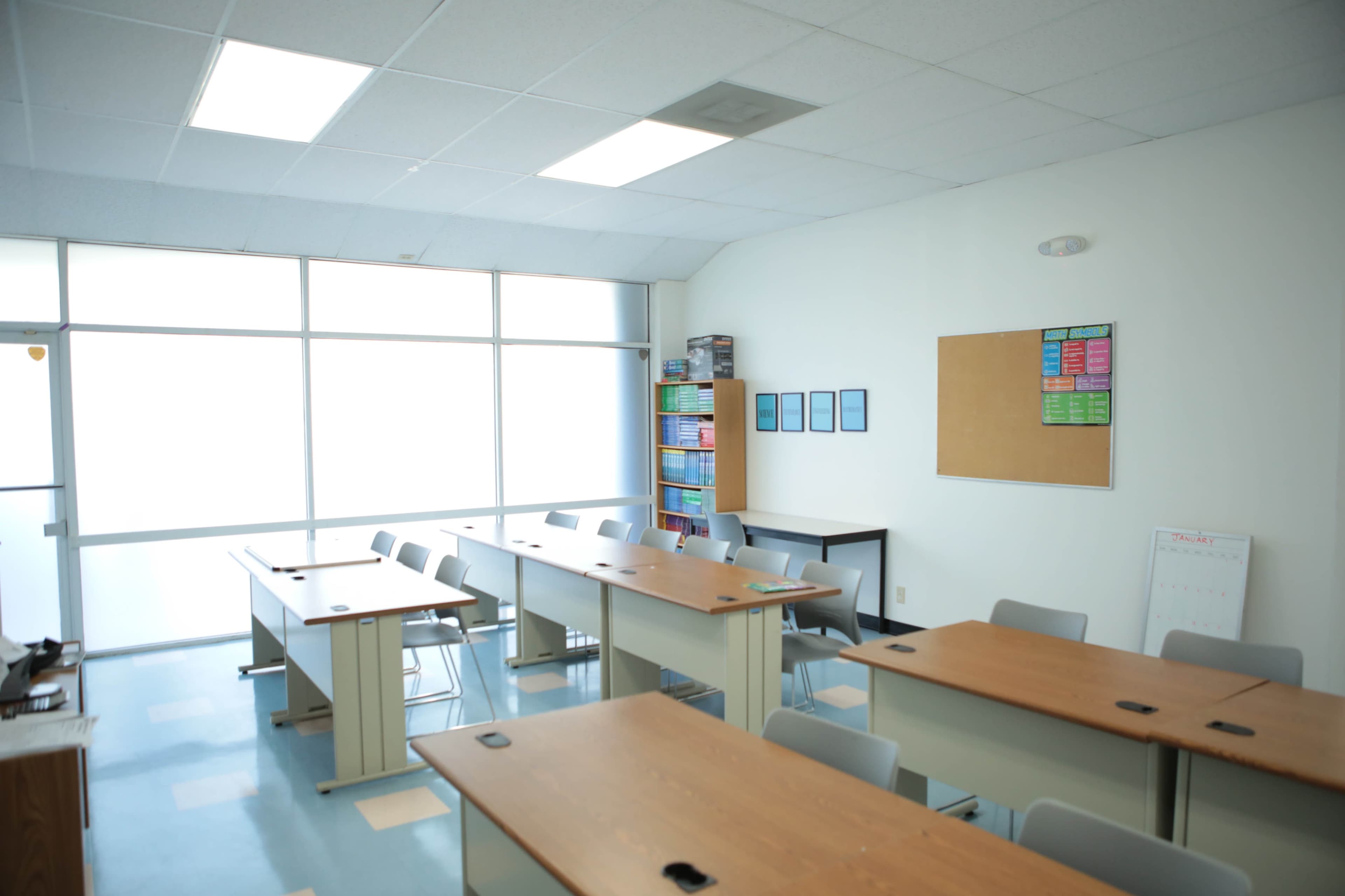 A classroom is arranged with several tables and chairs, featuring a whiteboard, a bulletin board, and shelves filled with books.