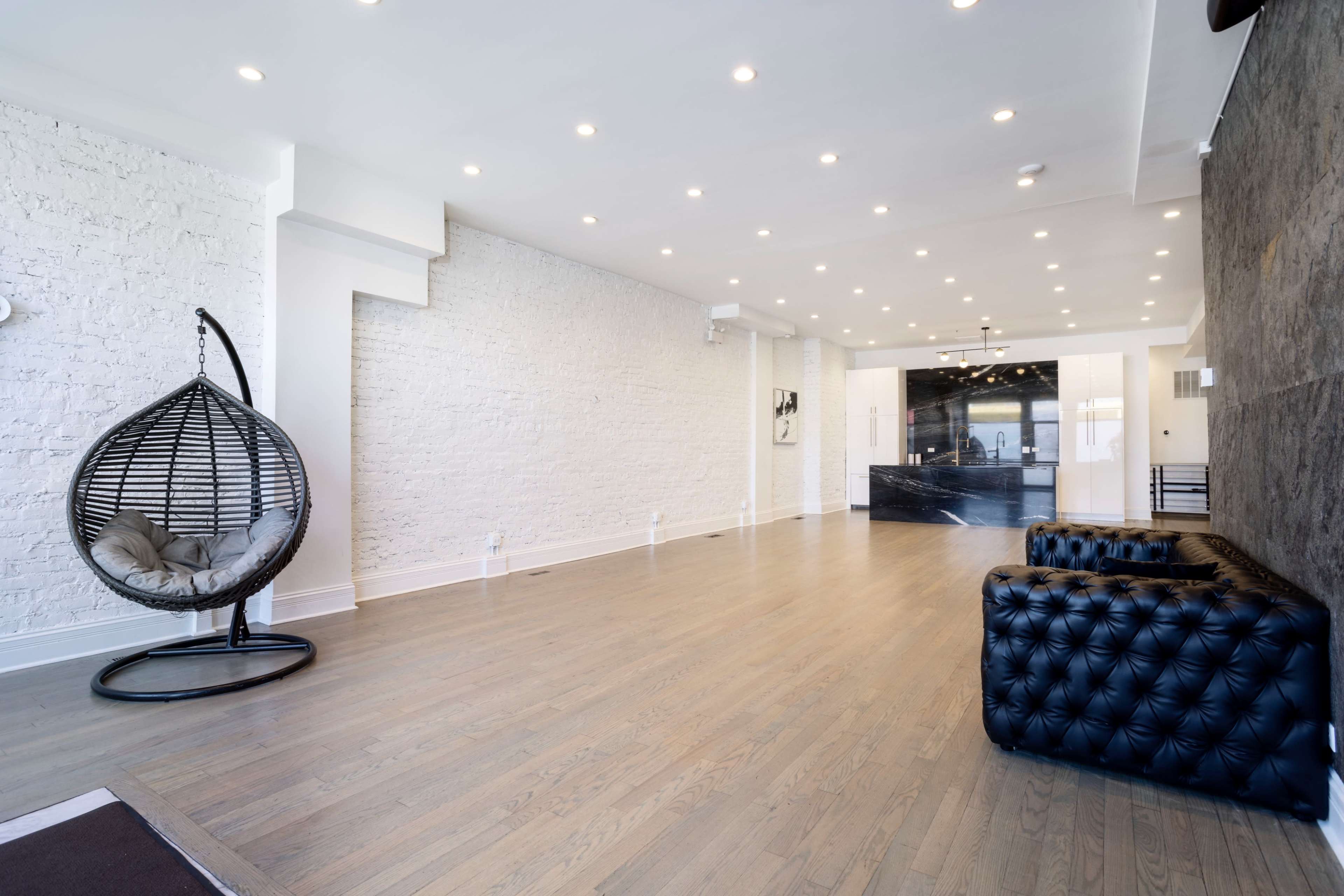 The image shows a spacious, modern lobby with a hanging chair on one side and a black tufted sofa on the other, featuring white walls and wooden flooring.