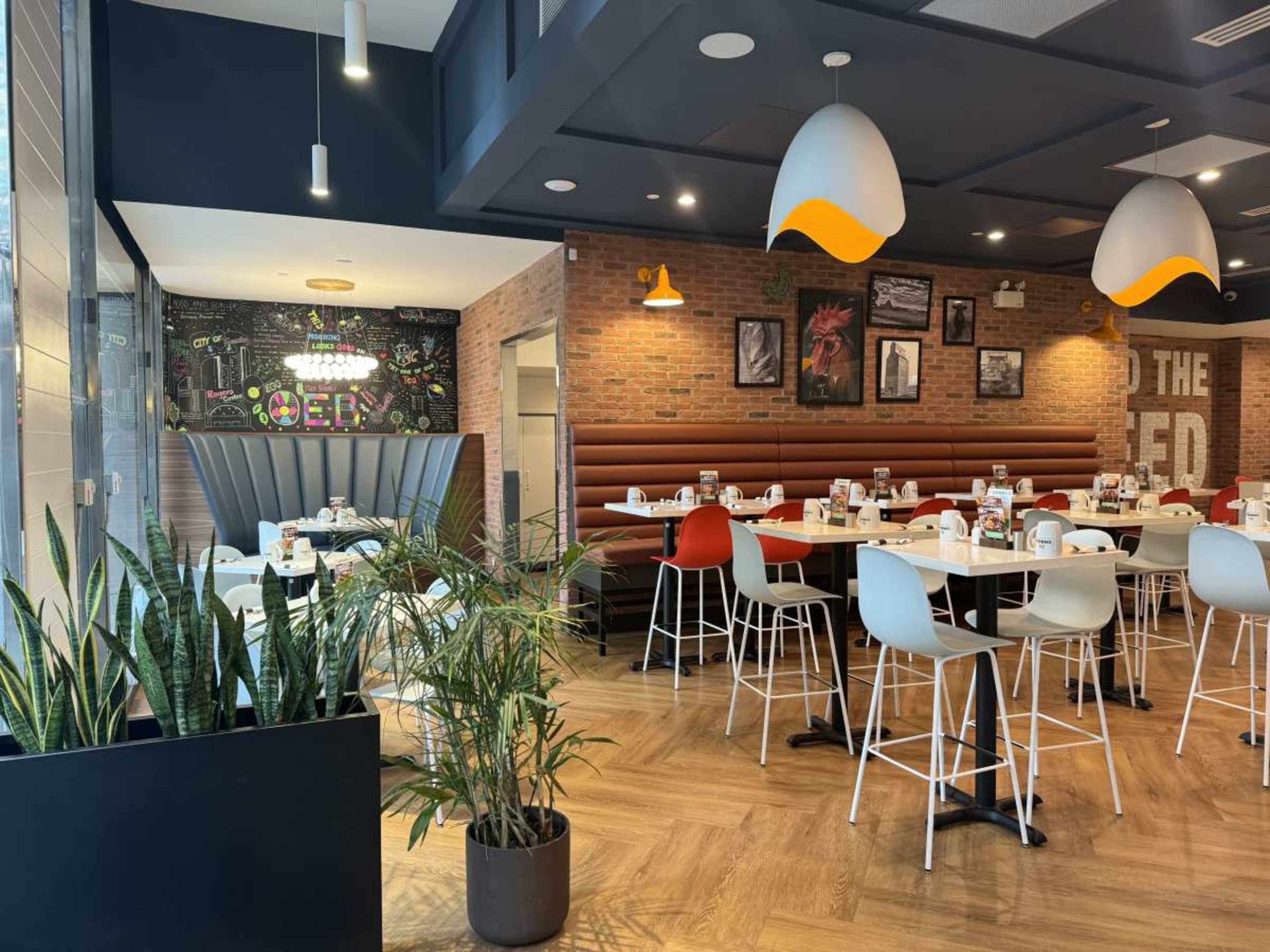The image shows a modern restaurant interior featuring wooden flooring, white tabletops, and colorful pendant lights hanging from the ceiling, with booth seating and decorative wall art.