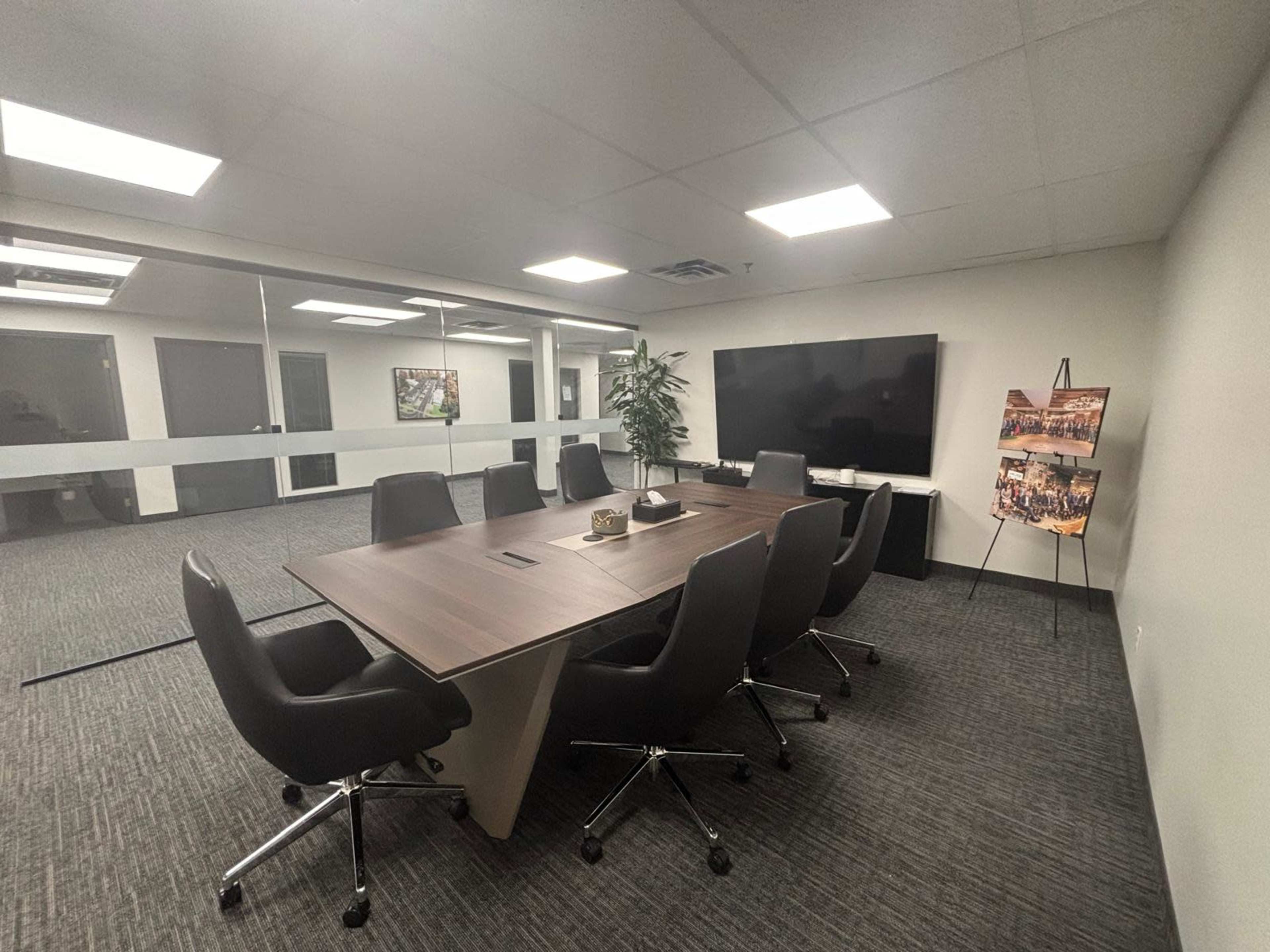 The image shows a modern conference room with a long table, black chairs, and a large screen on one wall.