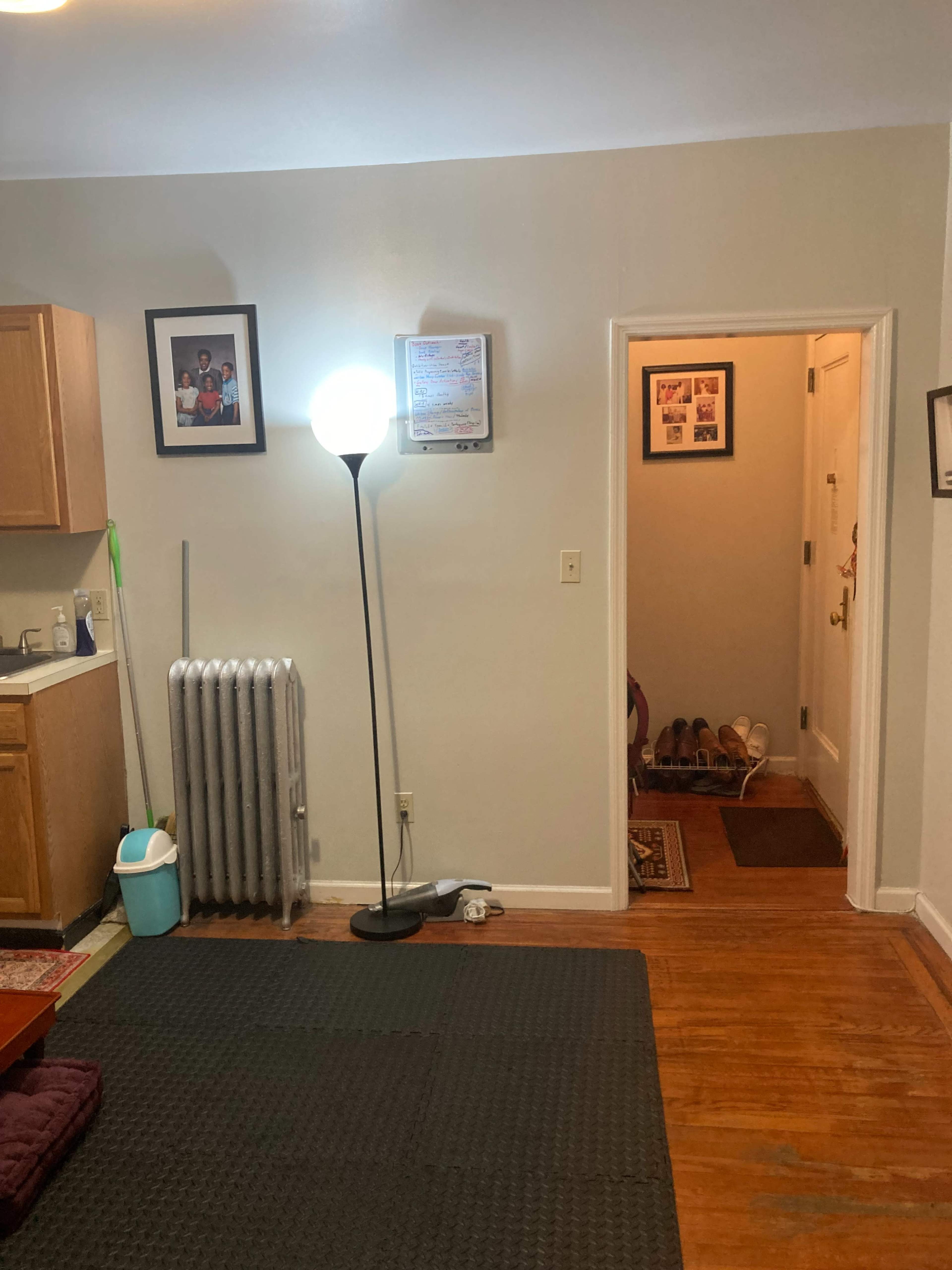 The image shows a room with a kitchen area on the left, a standing lamp in the center, and a doorway leading to a hallway on the right.