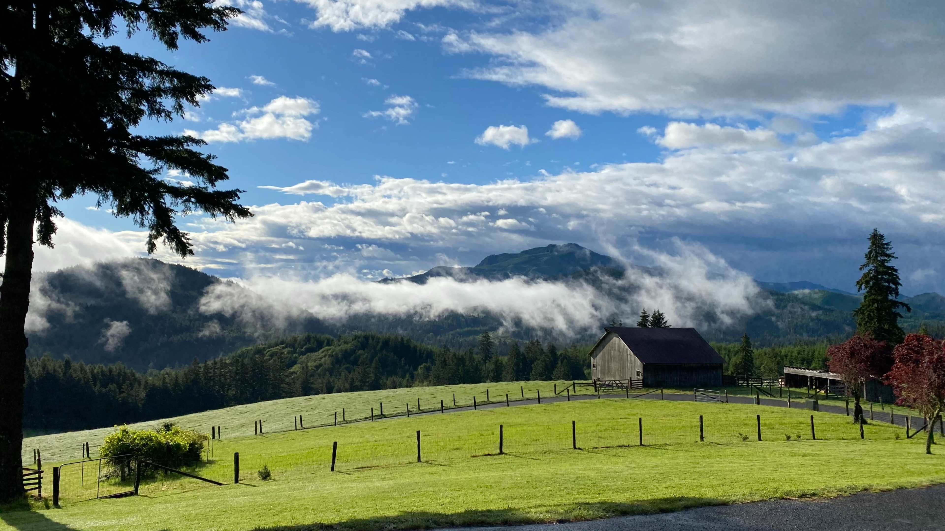 A wooden barn sits on a green hillside under a blue sky with scattered clouds and mist rolling over the mountains in the background.