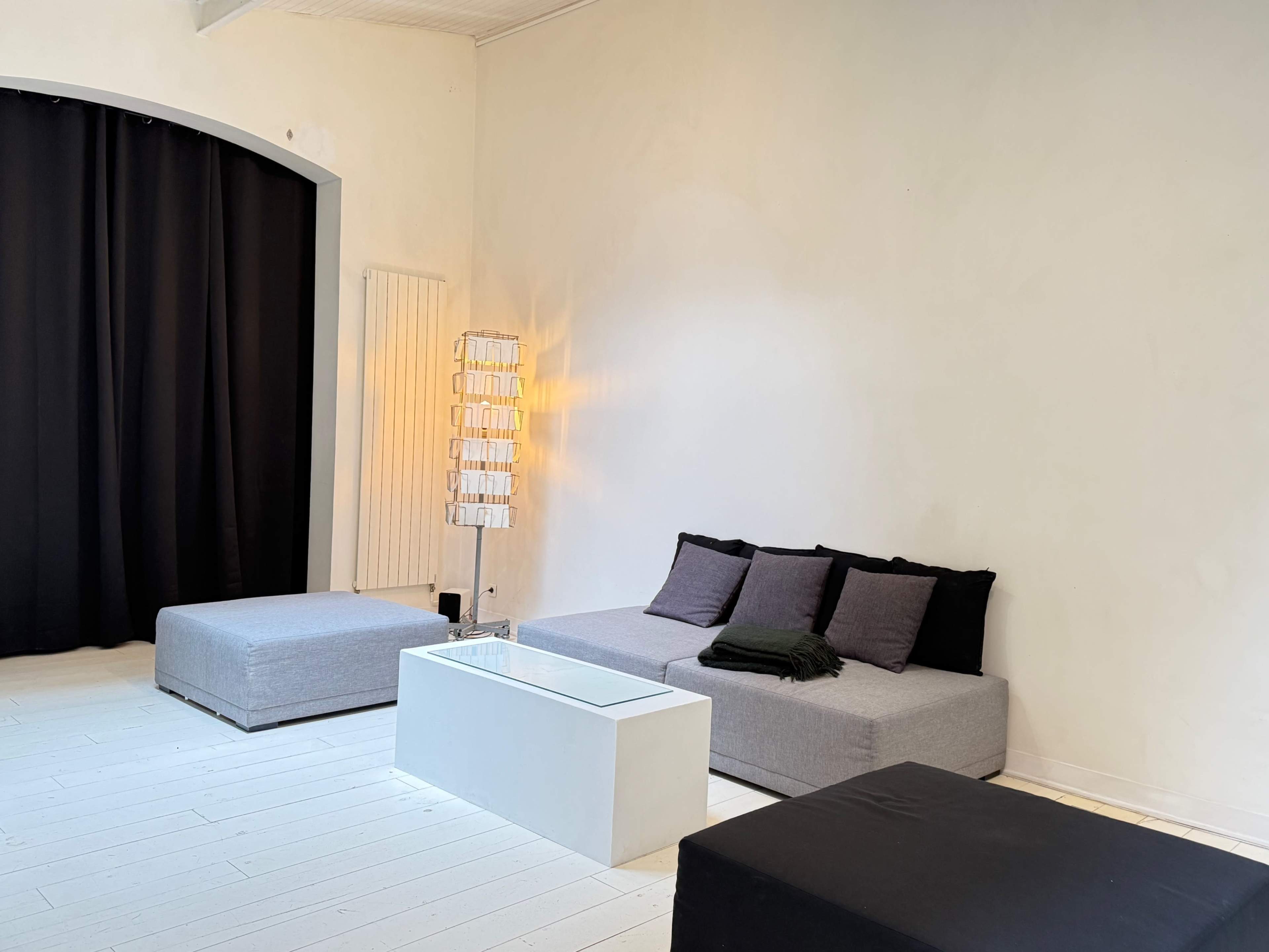The room features a minimalistic design with a gray sofa, a glass coffee table, and a floor lamp against a white wall, complemented by black curtains.