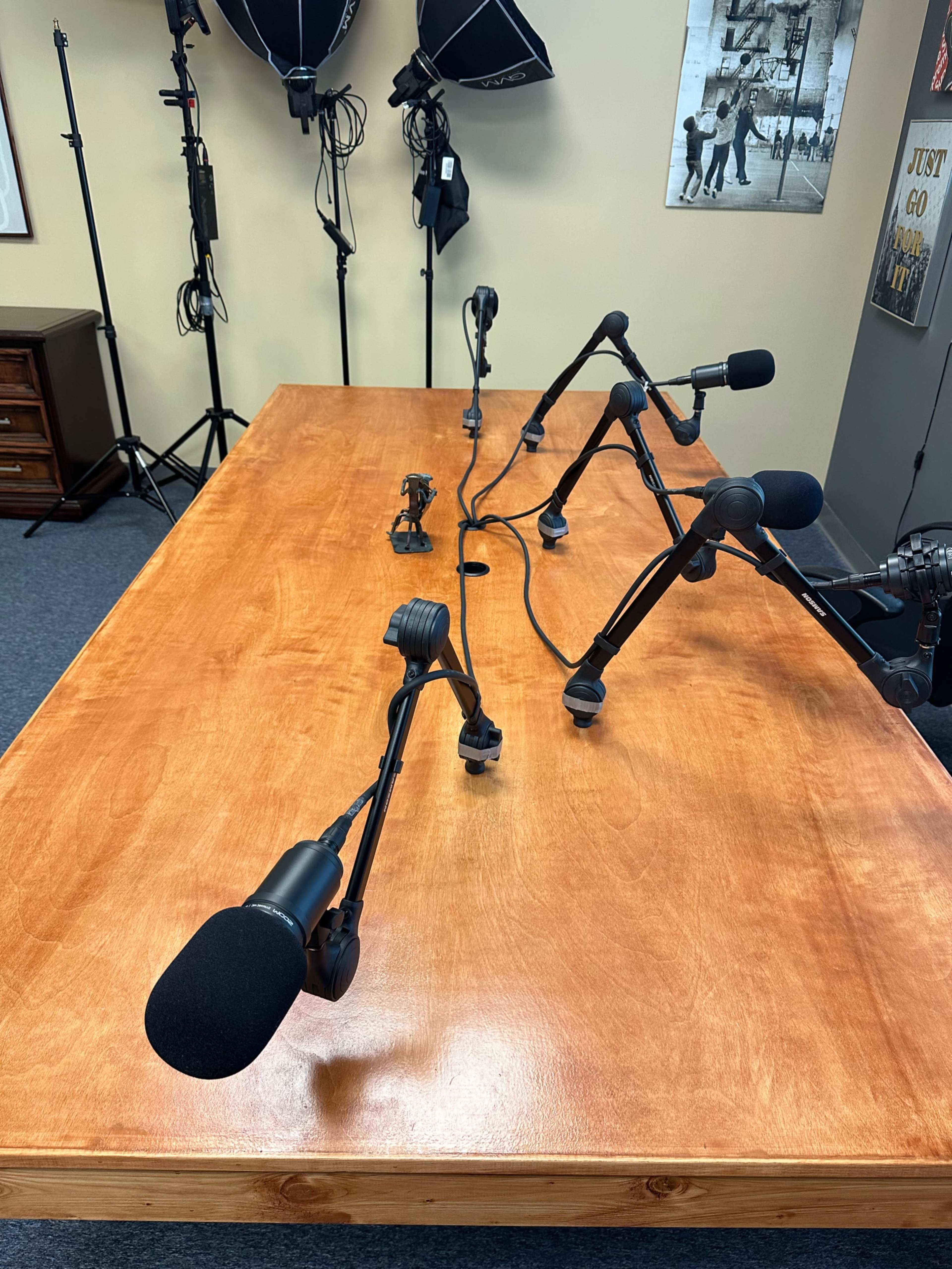 A wooden table is set up with multiple microphone stands arranged on top, surrounded by studio lighting equipment in a recording space.