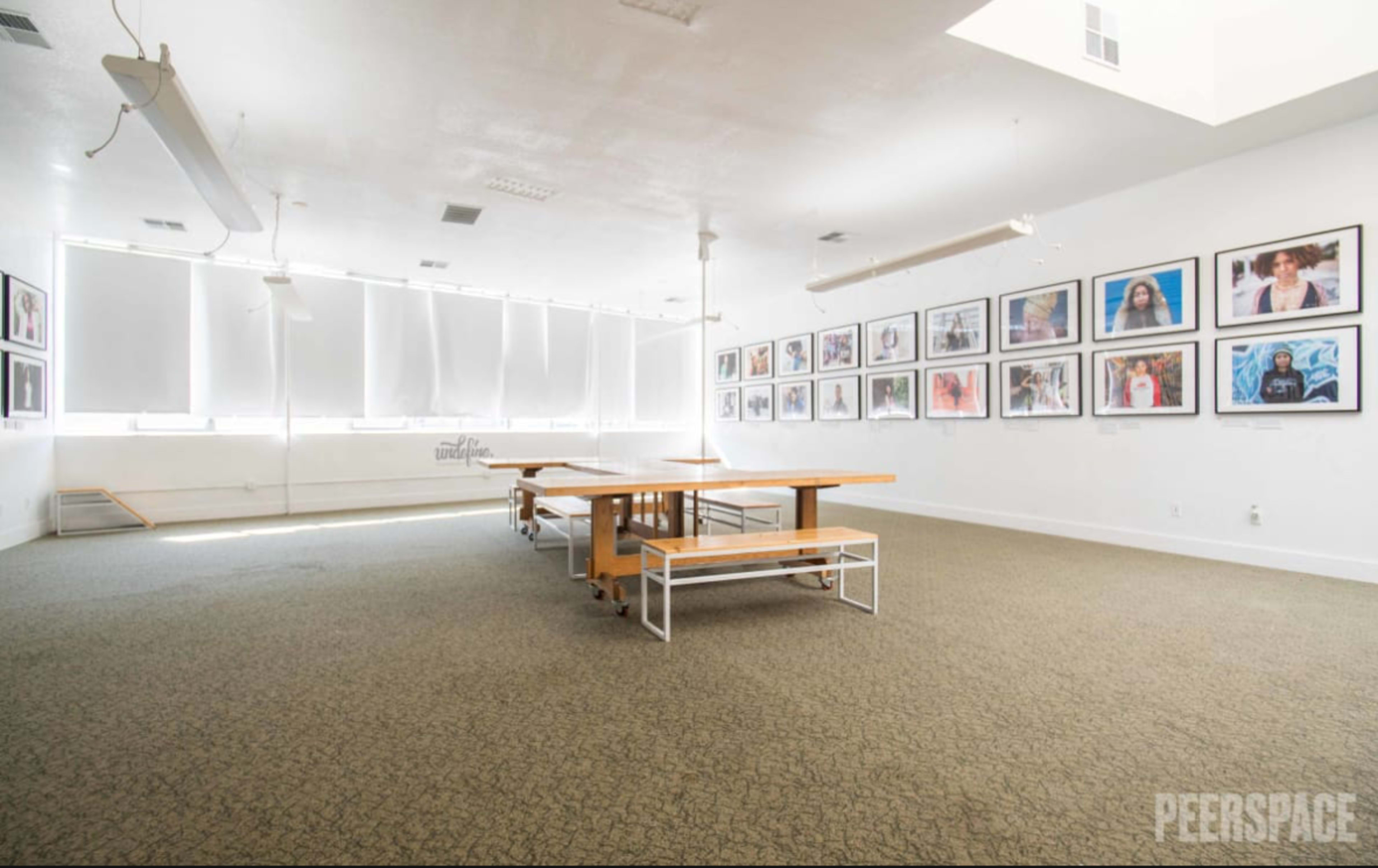 The image shows a spacious room with a long wooden table in the center, surrounded by white walls adorned with framed photographs, and large windows covered with sheer blinds.