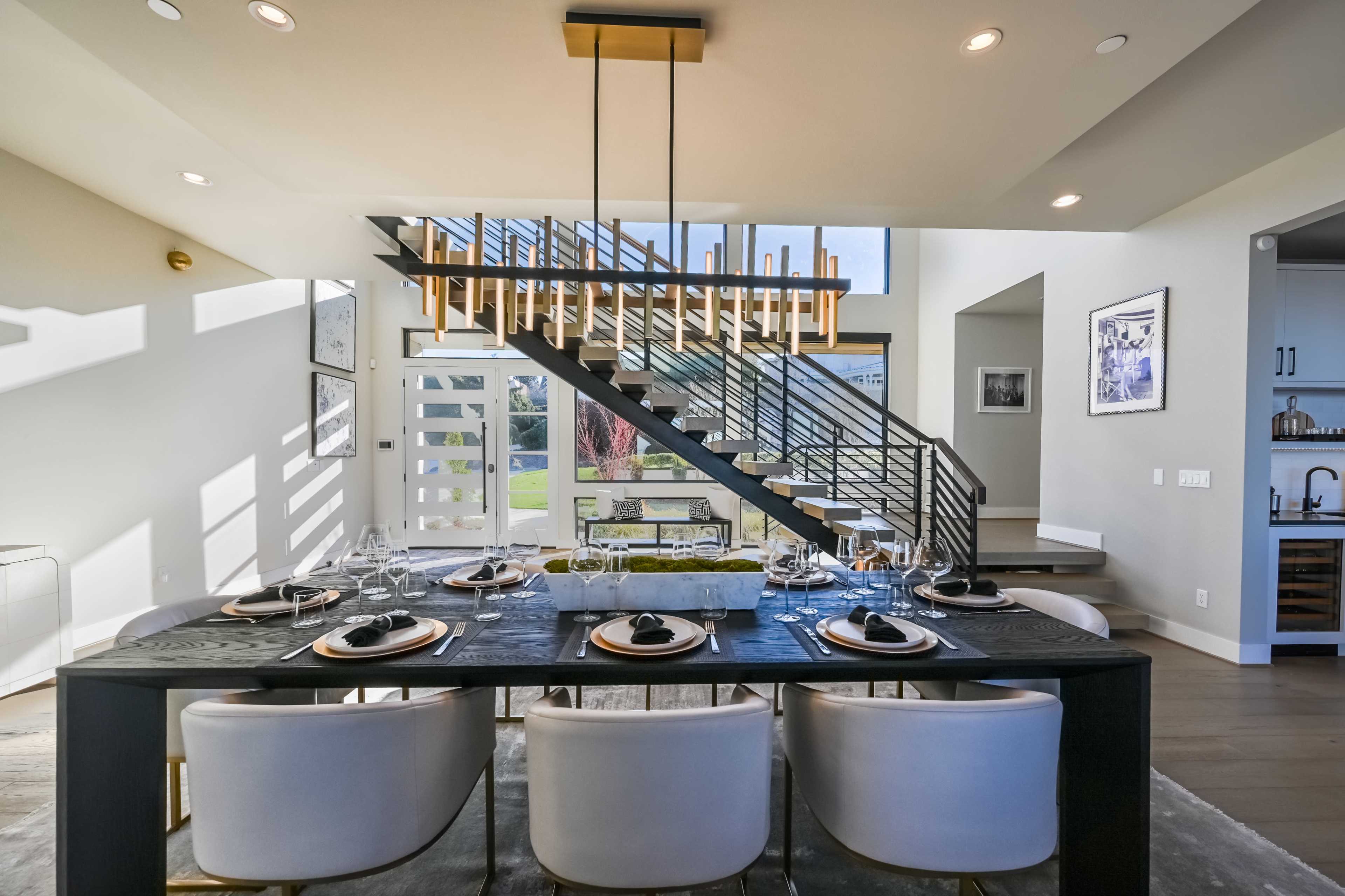 A modern dining area features a long black table set with elegant dishes, overlooking a staircase with a striking light fixture.