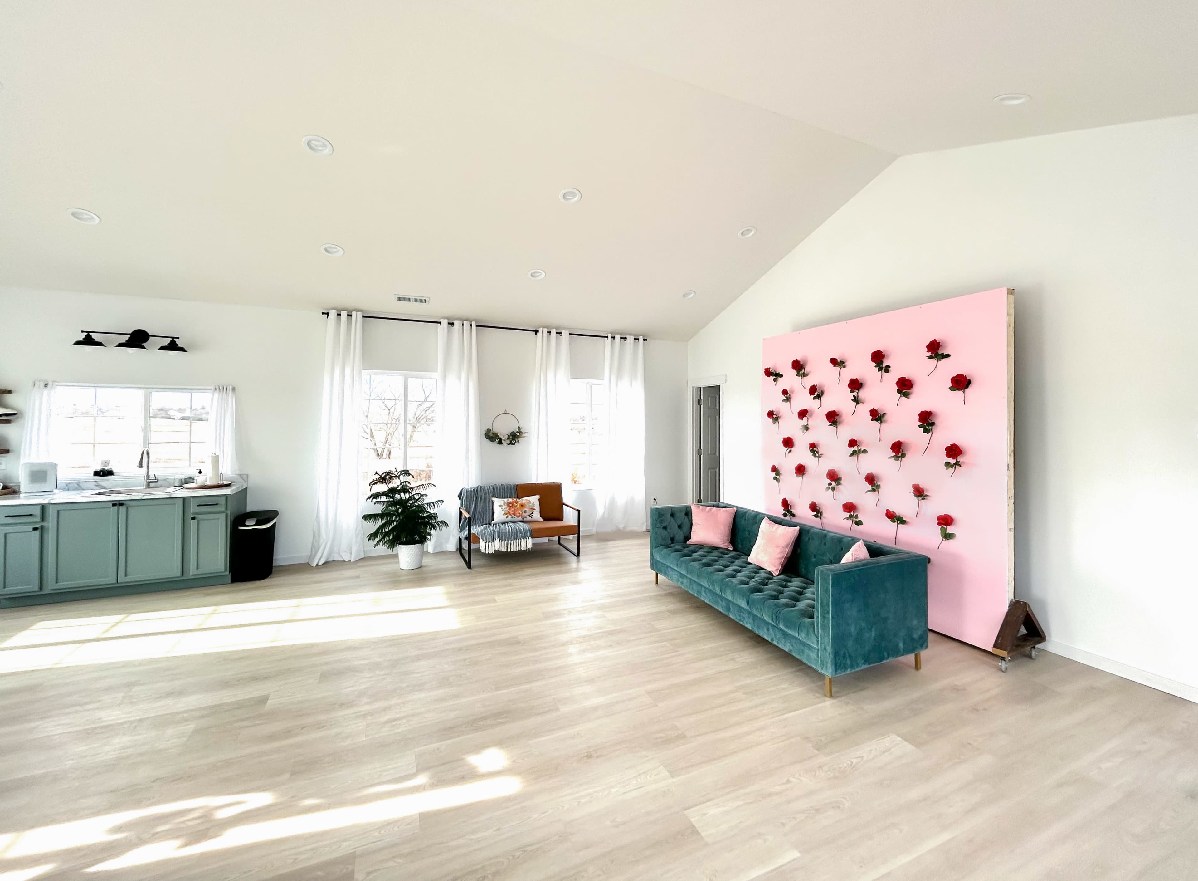 The bright interior features a green velvet sofa, a pink wall adorned with flowers, and a kitchenette area with large windows.