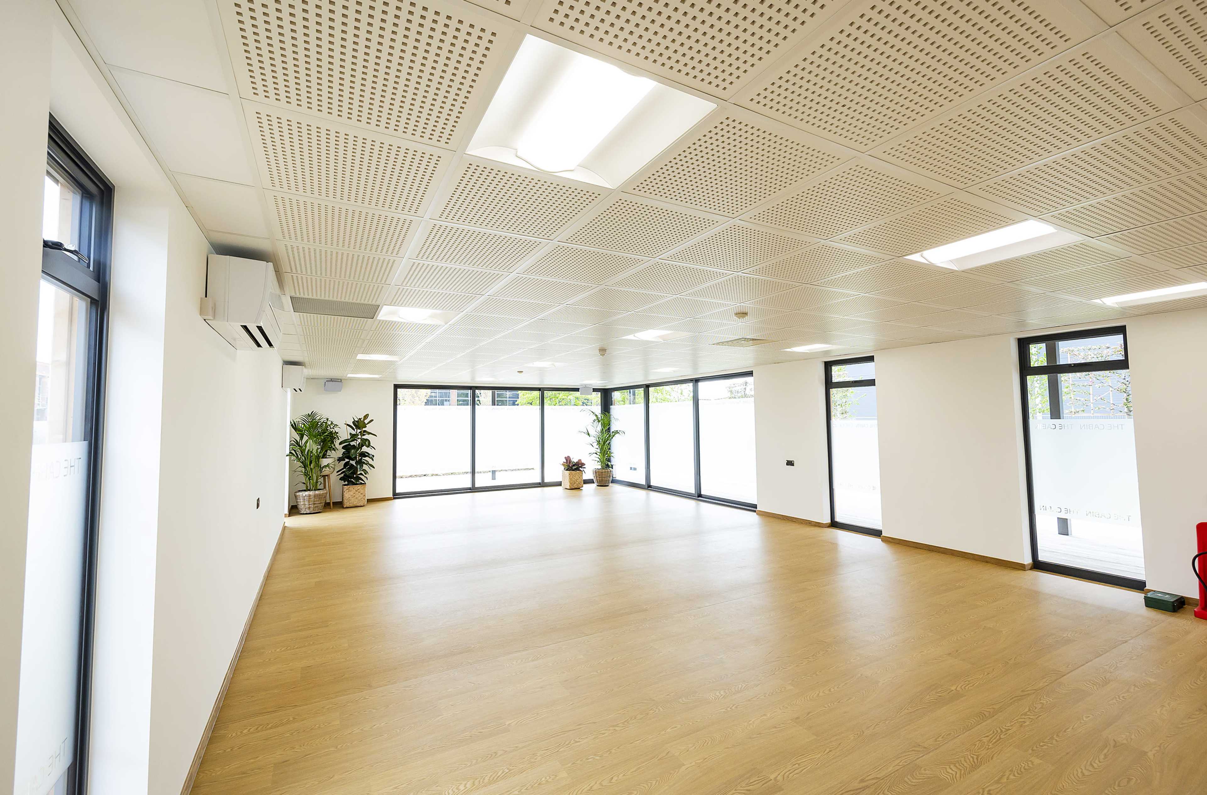 An empty office space with wooden flooring and large windows, featuring potted plants along the walls.