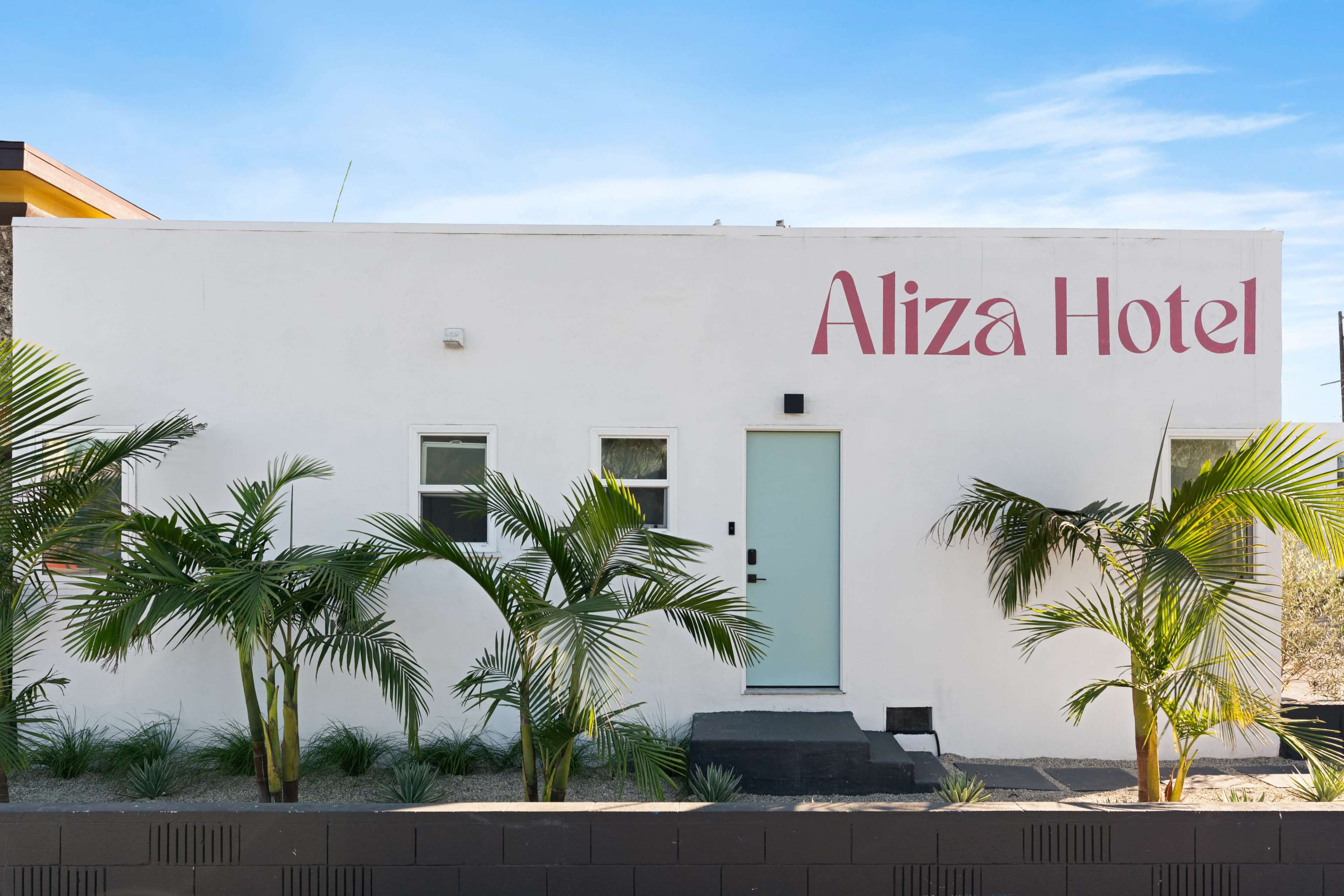 The image shows a white hotel facade with the text "Aliza Hotel" prominently displayed, flanked by palm plants and a light blue door.