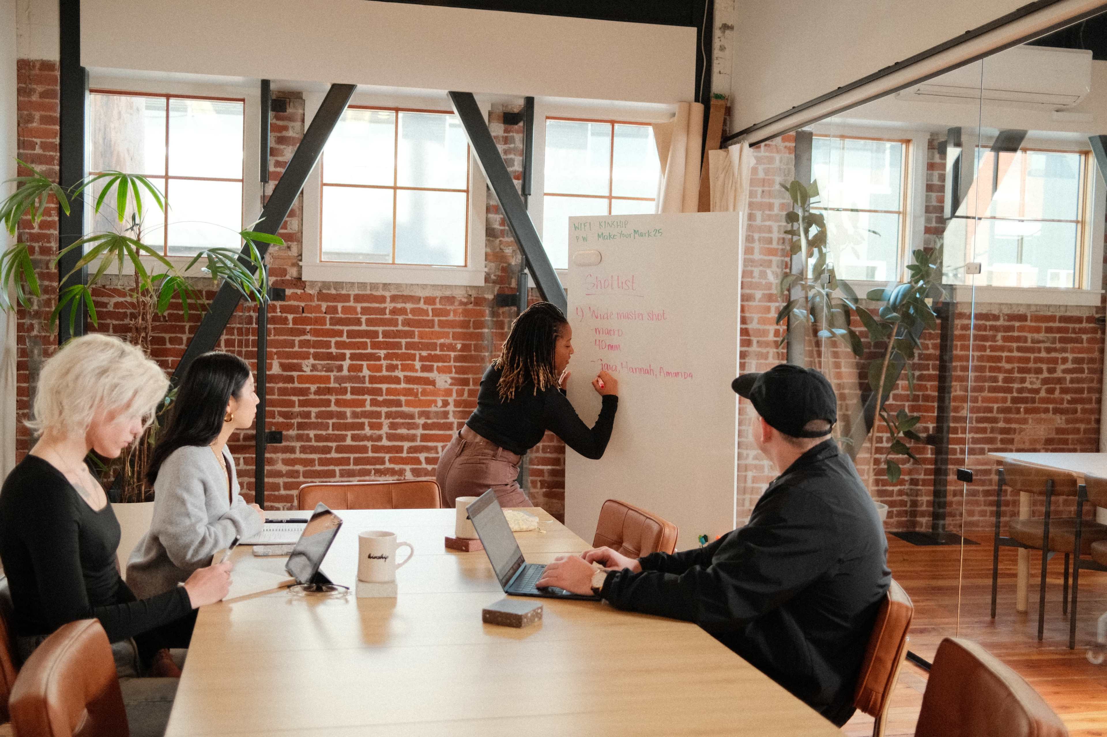 A group of four people are seated around a large table in a modern meeting room while one person writes on a whiteboard.