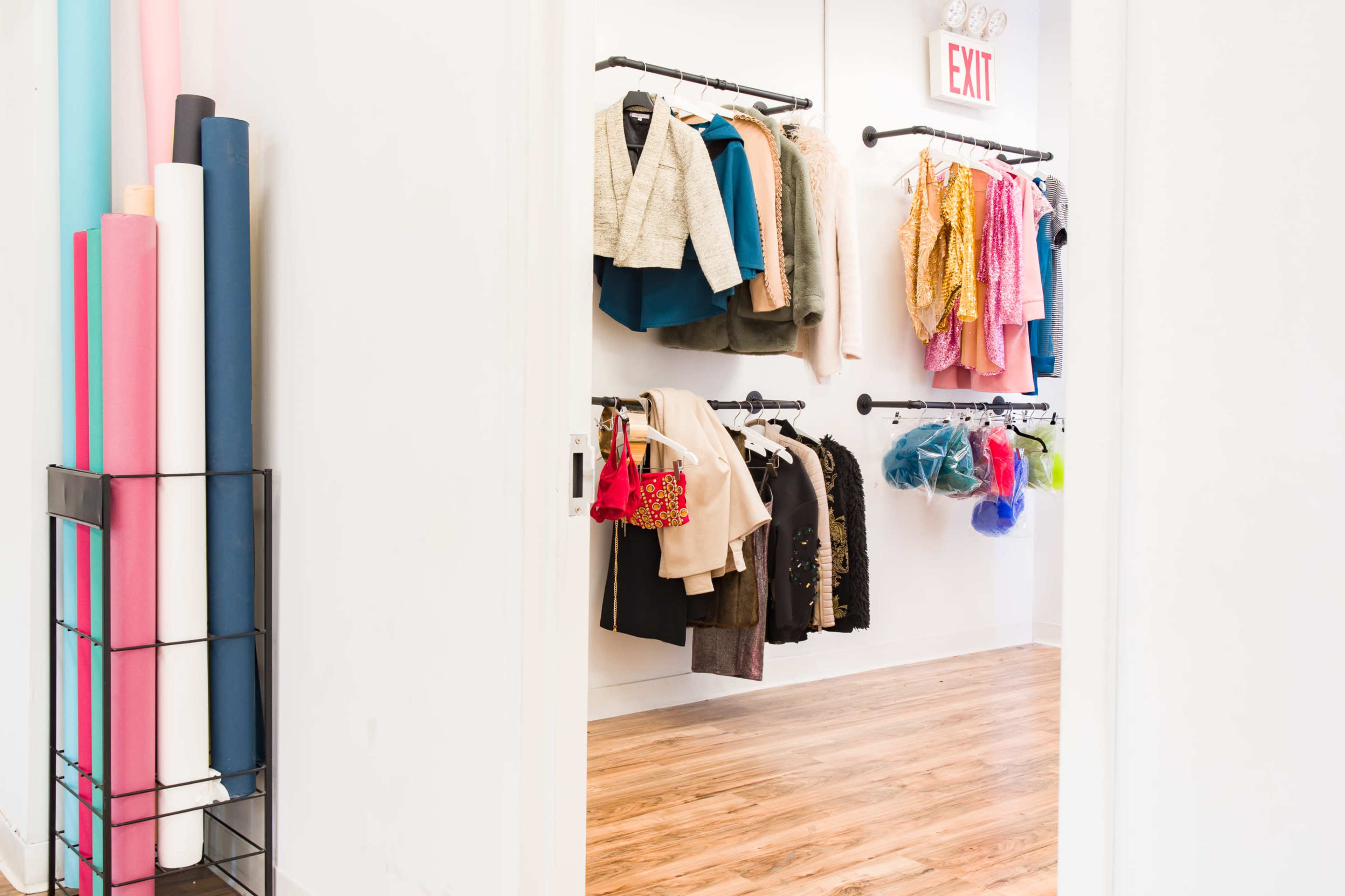 A clothing store displays a variety of coats and colorful accessories hanging on racks along one wall, with rolls of paper in different colors stored nearby.