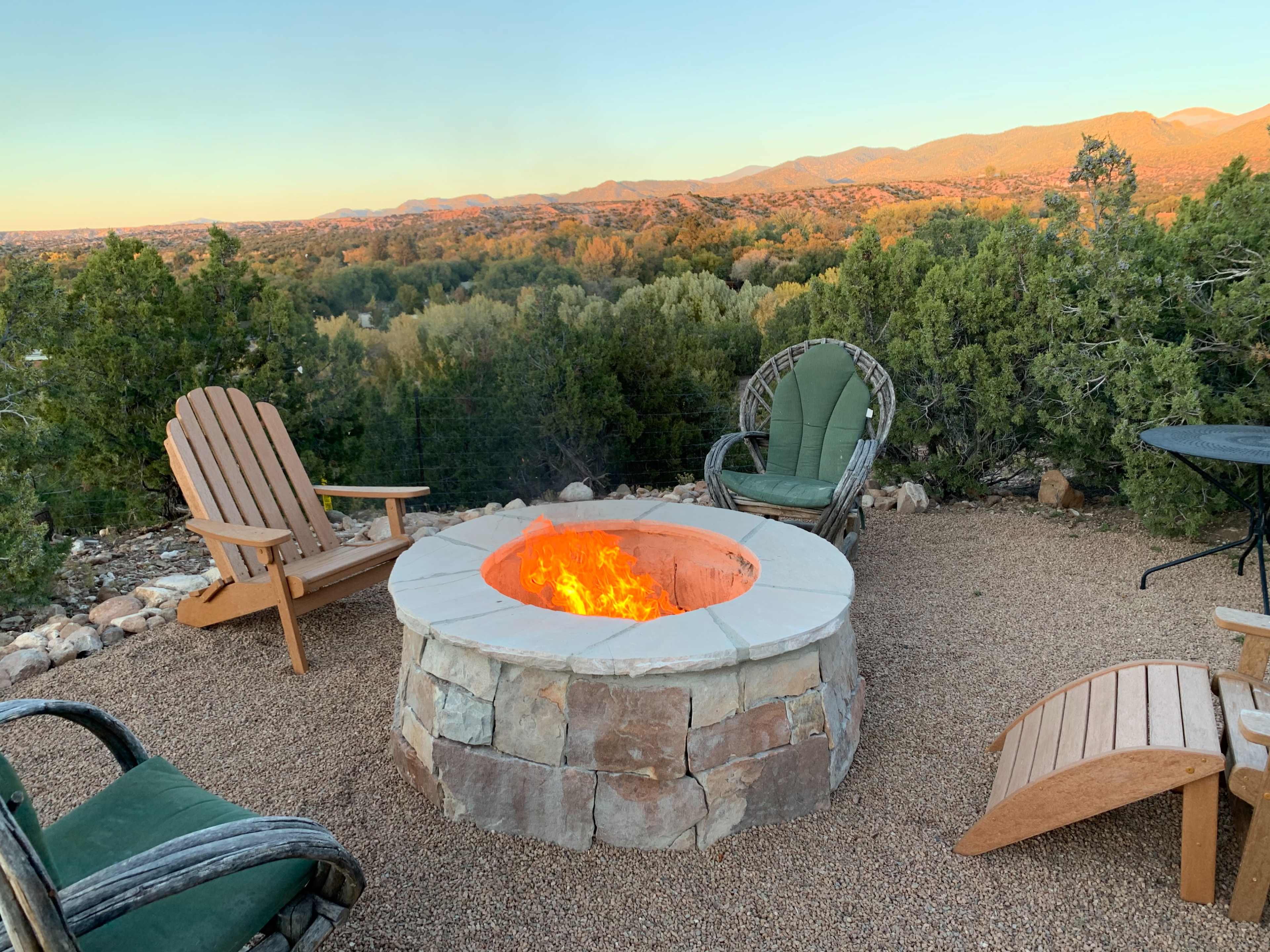 A stone fire pit with flames is surrounded by wooden and metal chairs, overlooking a landscape of trees and mountains at sunset.