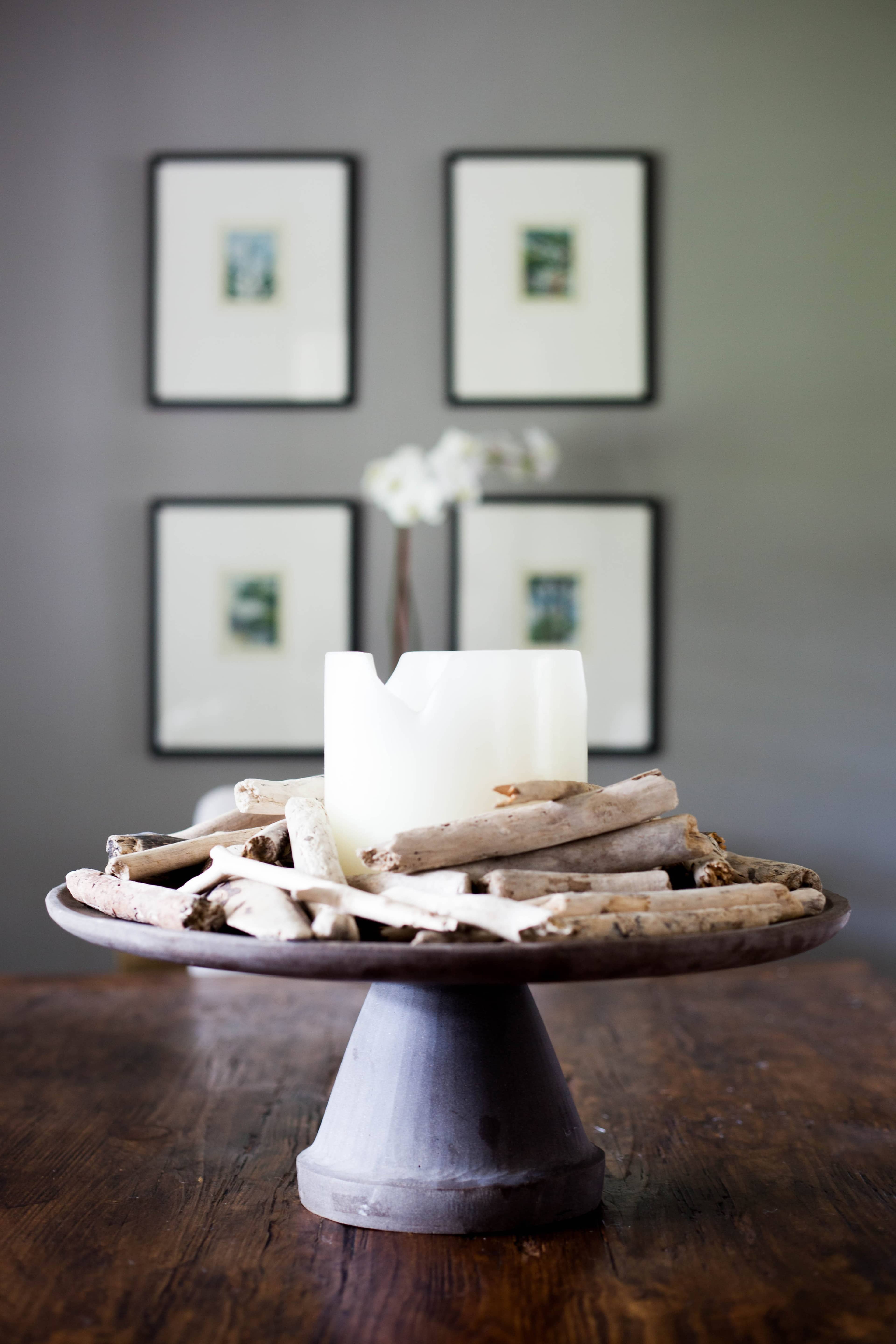 A white candle is surrounded by driftwood on a round pedestal, with framed pictures hanging on the gray wall behind it.