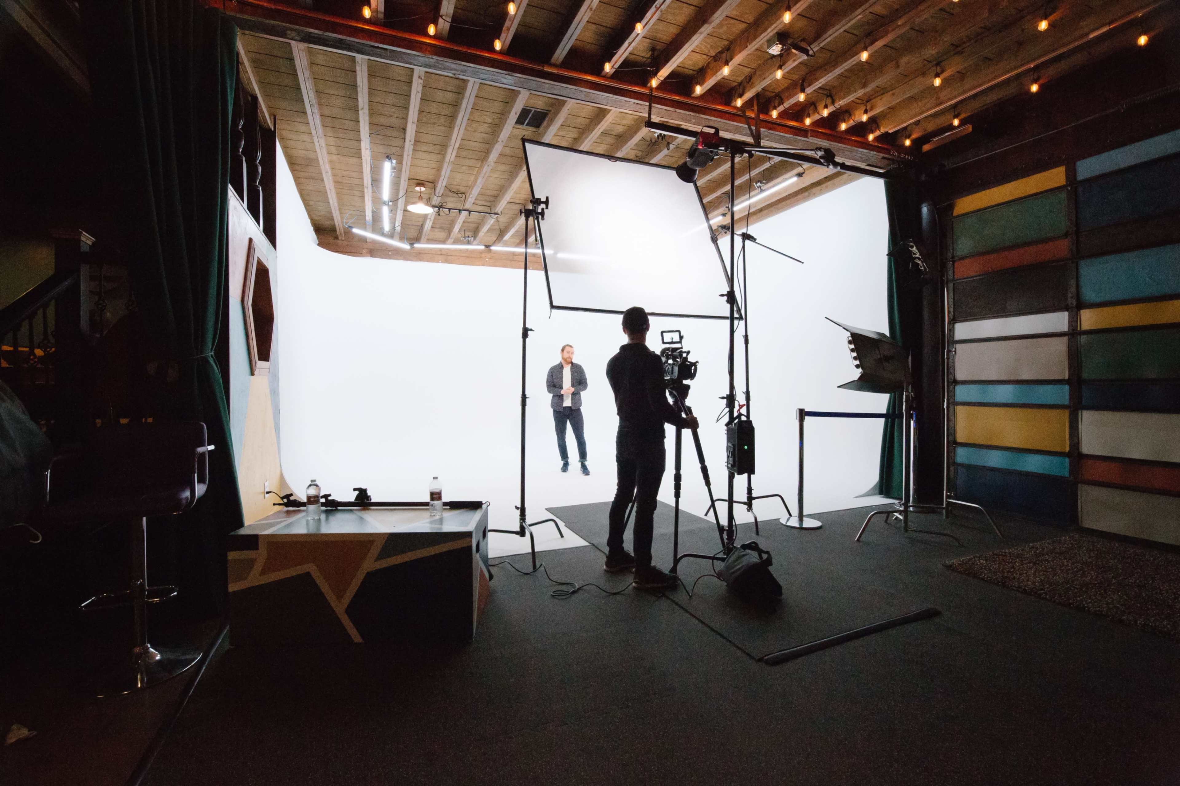 A person stands on a white backdrop inside a studio, while someone else sets up a camera and lighting equipment.